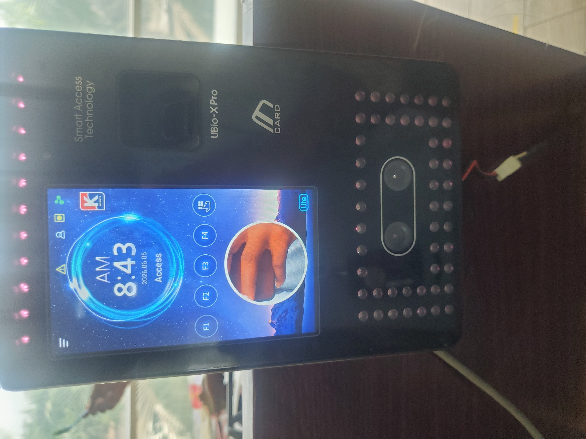 A black biometric access control device, identified by the text "Smart Access Technology" and "UBio-X Pro," is displayed prominently in a vertical orientation. The device features a large, active screen on its left side and a panel with numerous small, illuminated circular lights on its right. A white wire with red and black strands is visible connecting to the top right edge of the device, and a light-colored cable extends from its bottom right.
The device's screen shows "AM 8:43" and the date "2026.06.05," along with the word "Access." A circular image in the center of the screen displays a close-up of human fingers, suggesting an ongoing or recently completed hand or fingerprint biometric scan. Various interface elements are visible, including function keys labeled F1, F2, F3, F4, and icons representing Wi-Fi, user profiles, settings, messages, and door control. A "K SECURITY" logo and the text "Lite" are also present on the screen, along with a warning triangle icon and a hamburger menu icon. The device also displays "CARD" next to a card symbol on its upper body.
The scene depicts an indoor setting, likely an office or facility in Gwagwada, Nigeria, where the device is in operation. The device rests against a dark surface, and the background shows blurred details, possibly a room interior with a faint reflection of outdoor greenery through a window. The specific activity is the device displaying its operational status and a biometric scanning prompt. No discernible emotions are visible.