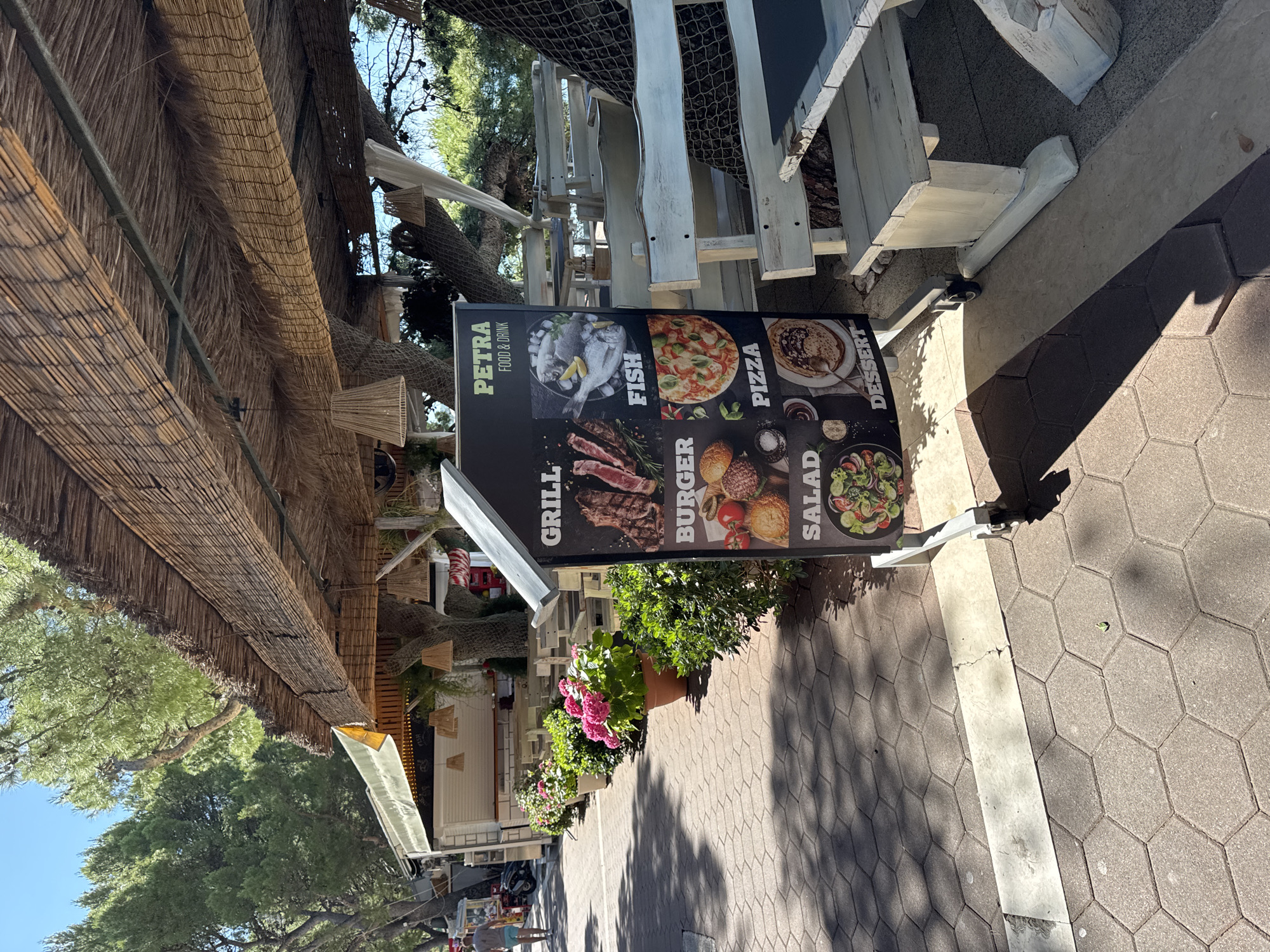 The image shows an outdoor dining area with a menu board in the foreground. The menu board advertises "PETRA FOOD & DRINK" and displays images of "GRILL," "FISH," "BURGER," "PIZZA," "SALAD," and "DESSERT." To the right of the menu board is a rustic wooden picnic table with white benches. In the background, there are trees, a building that appears to be a food stand or restaurant, and more outdoor seating. The ground is paved with hexagonal tiles. The overall scene suggests a casual dining establishment in a sunny, outdoor location. The presence of hydrangeas in pots and lush greenery indicates a pleasant, possibly seaside or park-like environment.