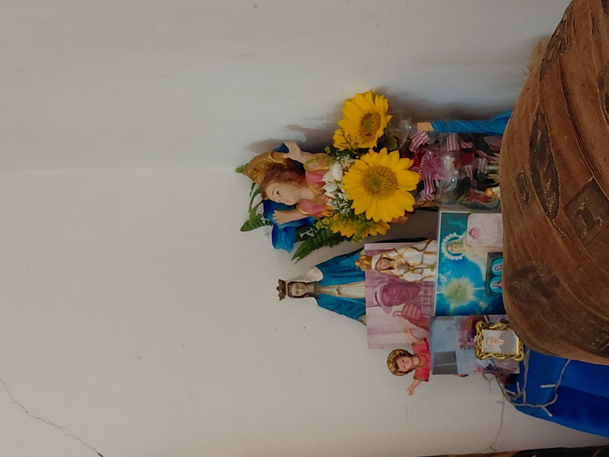 A domestic shrine is depicted in the corner of a room in Unknown, Unknown. The shrine is arranged on a surface draped with a blue fabric, partially obscured in the foreground by a large, textured brown object with prominent leaf-like patterns. Against a white wall, a collection of religious figures, photographs, and floral arrangements are displayed.
A crowned figurine of the Virgin Mary, dressed in a blue mantle over a white garment, stands on a rectangular base. To its lower left, a small figurine, possibly the Infant Jesus, with outstretched arms and wearing red and gold attire, is visible. To the right and slightly behind the main Marian figure, partially obscured by blue and yellow artificial flowers, is a child-angel figurine in pink and gold with outstretched arms. A smaller crowned Marian figure in white and gold stands to the lower right of the larger Marian figure, adjacent to a photographic print depicting a man in a hat giving a thumbs-up. To the right of the man's print, a depiction of the Virgin Mary with a radiating halo against a blue background is visible.
A bouquet of yellow sunflowers, accompanied by white and green filler flowers, is arranged in a clear glass vase. The vase contains multiple small, folded red, white, and blue striped flag-like items. A small, ornate gold-framed photograph of an older man with a mustache, wearing a suit, is placed on the blue fabric in the lower left portion of the altar. The white wall behind the display exhibits a prominent vertical crack extending from the upper left and minor surface imperfections.