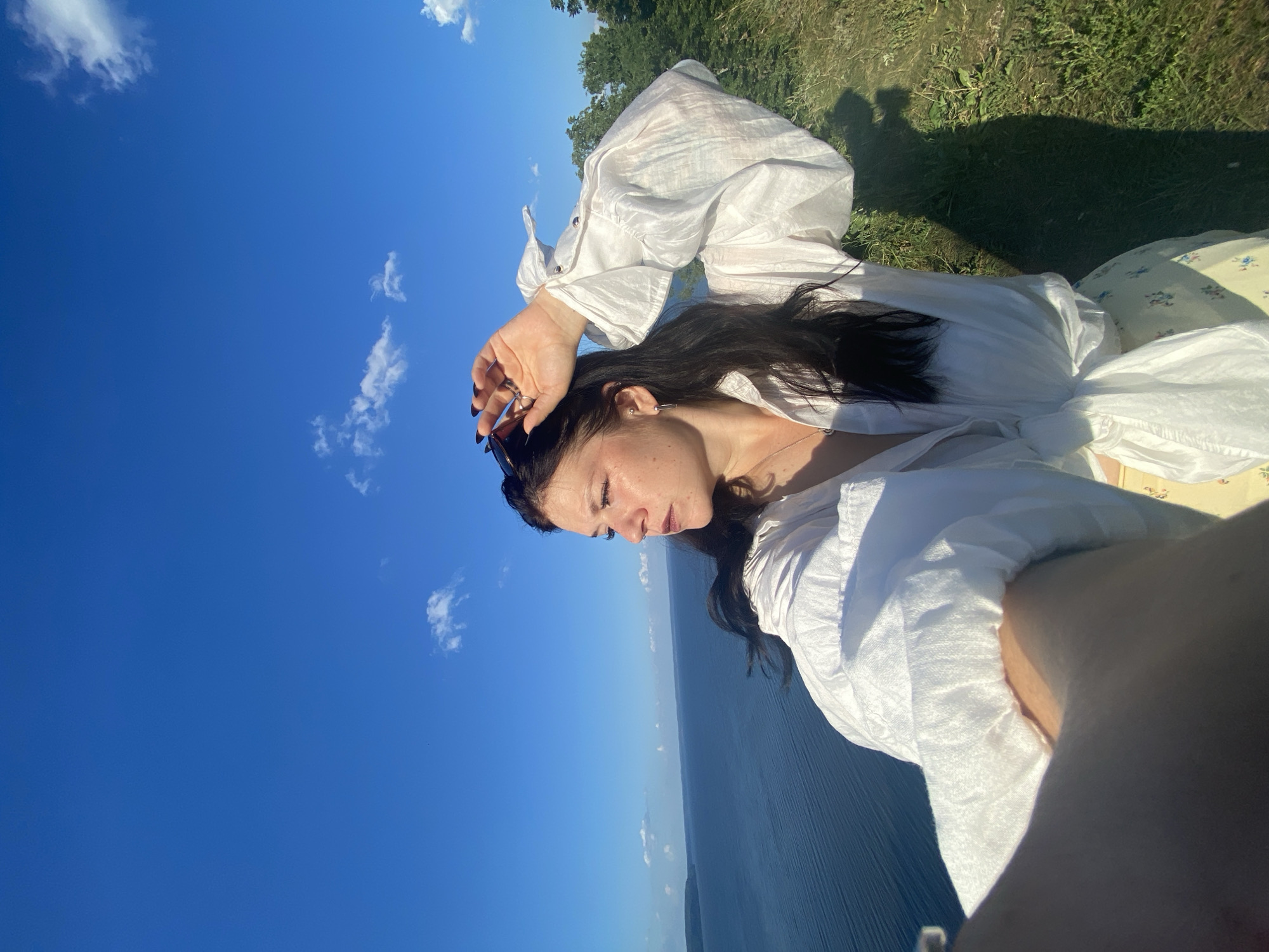 A young woman with dark hair, wearing a white ruffled shirt and a floral skirt, stands on a grassy cliff overlooking a wide, calm body of water under a bright blue sky with scattered white clouds. She is looking to her left, with her right hand raised to shade her eyes from the sun, holding a pair of sunglasses. The scene is captured in a selfie-style, with a portion of the photographer's arm visible in the lower left corner. The location is the city of Kostovaty, Russia.