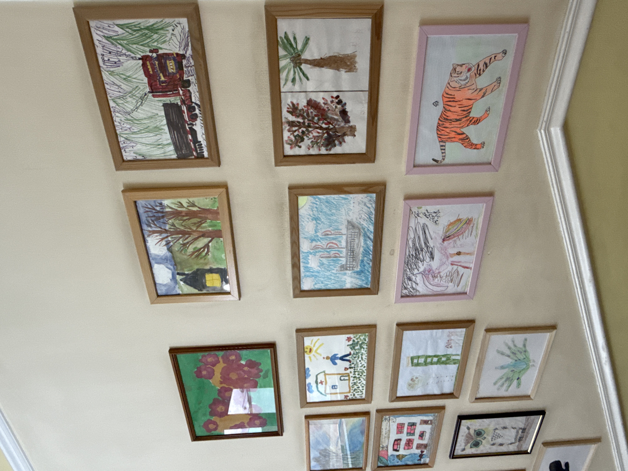 The image displays a collection of framed children's drawings hung on a pale yellow wall, likely in a residential setting. The drawings are diverse, featuring subjects such as a red truck, houses, trees, a ship, a person with a sun, an owl, abstract green shapes, and a tiger. Some frames are wooden, while others are pink or black. The wall is smooth and appears to be painted. A white molding runs along the bottom of the frame, suggesting the bottom of a staircase or a wall transition. There are no people visible in the image. The scene does not provide any specific information to indicate its location in Saint Petersburg, Russia.