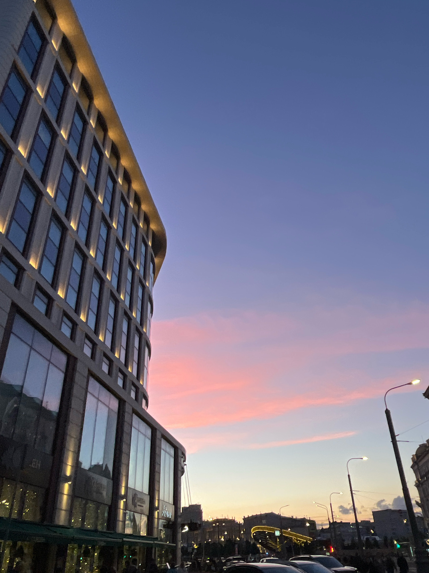 The image captures a twilight scene in Moscow, Russia, focusing on the exterior of a modern, curved building with a facade of large windows and illuminated horizontal bands along its upper floors. The building's architecture suggests a commercial or office space. The sky above transitions from a deep blue at the top to lighter shades of purple and pink near the horizon, indicating sunset. Wispy pink clouds are visible in the lower part of the sky.
In the foreground and to the right, a street scene unfolds. Several streetlights are present, one prominently in the right foreground with a curved arm. Cars are parked or driving on the street, partially visible. In the background, city buildings and structures, including a brightly lit bridge-like structure, are silhouetted against the fading light. A few indistinct figures of people can be seen on the sidewalk. The overall impression is of an urban environment at dusk, with the modern architecture contrasting with the natural beauty of the twilight sky.