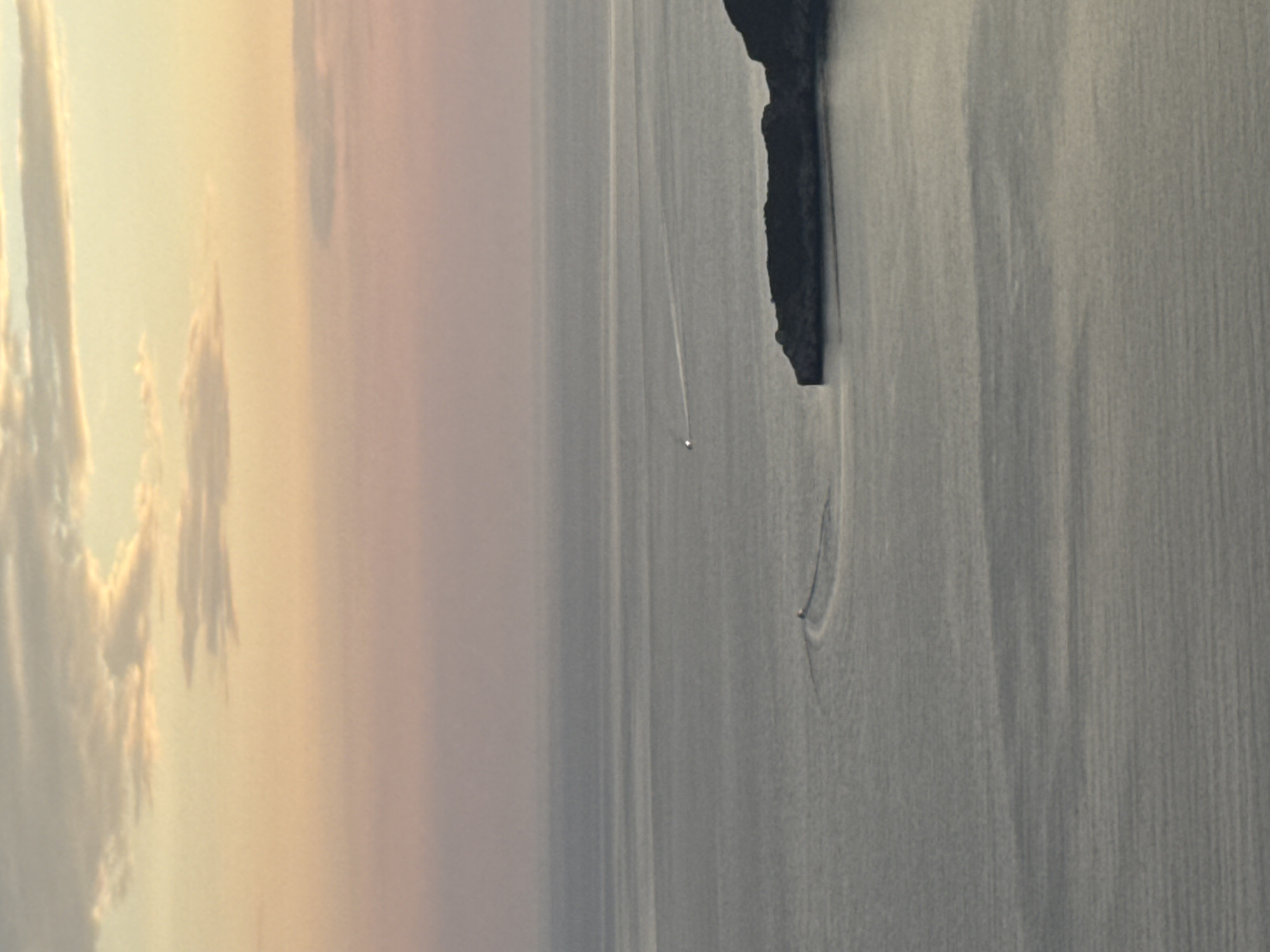 The image depicts a serene seascape with a boat traversing the calm waters. On the left, fluffy clouds are illuminated by the soft, warm glow of what appears to be sunrise or sunset. The colors in the sky transition from a pale yellow to a delicate pink and then to a muted grey, suggesting a tranquil atmosphere. The sea on the right is a uniform grey, reflecting the muted tones of the sky and creating a sense of peacefulness. A small, dark object, likely a boat or a buoy, is visible in the middle distance, leaving a subtle wake that ripples across the water's surface. To the far right, a dark, rocky outcrop juts out from the water, its textured surface providing a stark contrast to the smooth expanse of the sea. The overall scene evokes a sense of solitude and natural beauty, possibly captured during the early morning or late evening hours. The location context of Pržno, Montenegro, hints at a coastal setting with likely clear skies and calm seas typical of this region during favorable weather conditions. No visible text is present in the image.