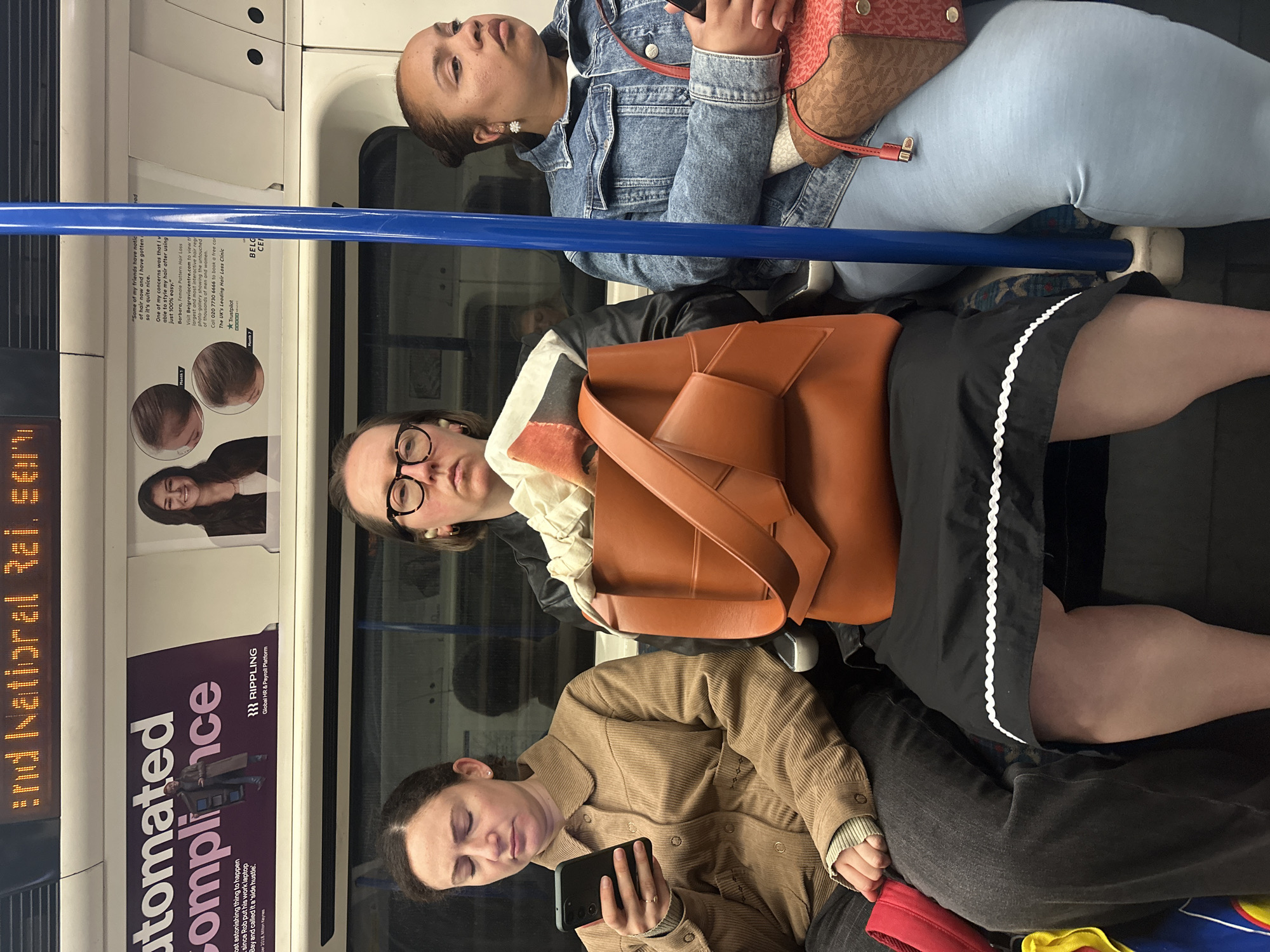 This photograph captures a moment on the London Underground, specifically in a train carriage. Three women are visible. The woman at the top is light-skinned and wearing a denim jacket and light blue jeans, with a small brown purse on her lap. The woman in the middle is light-skinned and wearing glasses, a brown handbag, a white top with a high frilled collar, and a dark skirt or dress. The woman at the bottom is light-skinned and looking at her phone, wearing a brown corduroy jacket and dark trousers.
Advertisements are visible on the train walls, including one for "Rippling" - a global HR and payroll platform, and another for a hair loss clinic. A blue handrail runs horizontally across the frame. A digital display on the train reads "and Natioral Rai ser," part of an incomplete message. The carriage appears to be relatively full, given the positioning of the passengers.