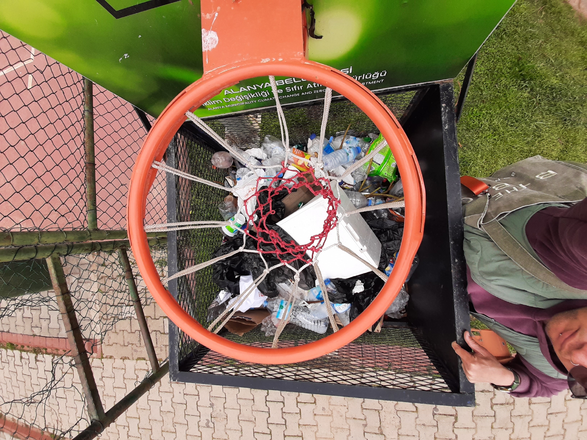 A top-down shot reveals an orange basketball hoop mounted over a black metal trash container. The container is filled with an assortment of trash, including plastic bottles and black plastic bags. The basketball hoop has a white rope net and a red twine net inside. To the left of the container is a chain-link fence on a red brick surface. To the right, part of a person is visible; they are wearing a green and grey vest and a maroon shirt, with a watch on their wrist. The background features green grass and a green sign that says "Alanya Belediyesi" (Alanya Municipality) along with "Klim Değişikliği ve Sıfır Atı" (Climate Change and Zero Waste) in Turkish. The text "ALANYA MUNICIPALITY CLIMATE CHANGE AND ZERO" is also present, indicating the location as Alanya, Turkey.