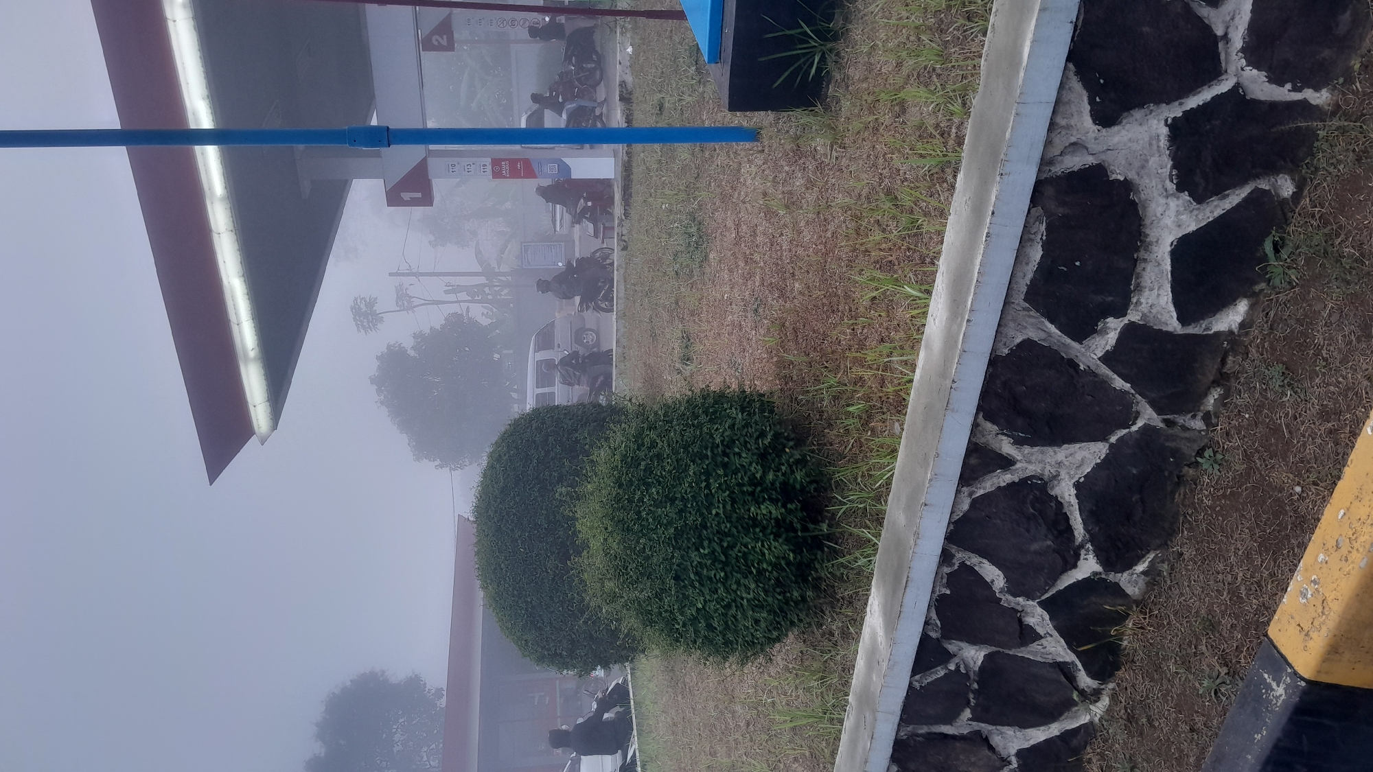 The image captures a foggy morning scene at a gas station in Subang, Indonesia. In the foreground, a stone wall separates a grassy area from a yellow and black striped barrier. Two neatly trimmed bushes are situated on the grassy area. Behind the bushes, several motorcycles and a small van are visible, with people on the motorcycles. The gas station canopy, painted red and white, extends over the pumps, which are partially obscured by the fog. A tall blue pole stands near the pumps, and signs with numbers like "1" and "2" are visible. The overall atmosphere is hazy due to the fog, creating a muted and subdued visual.