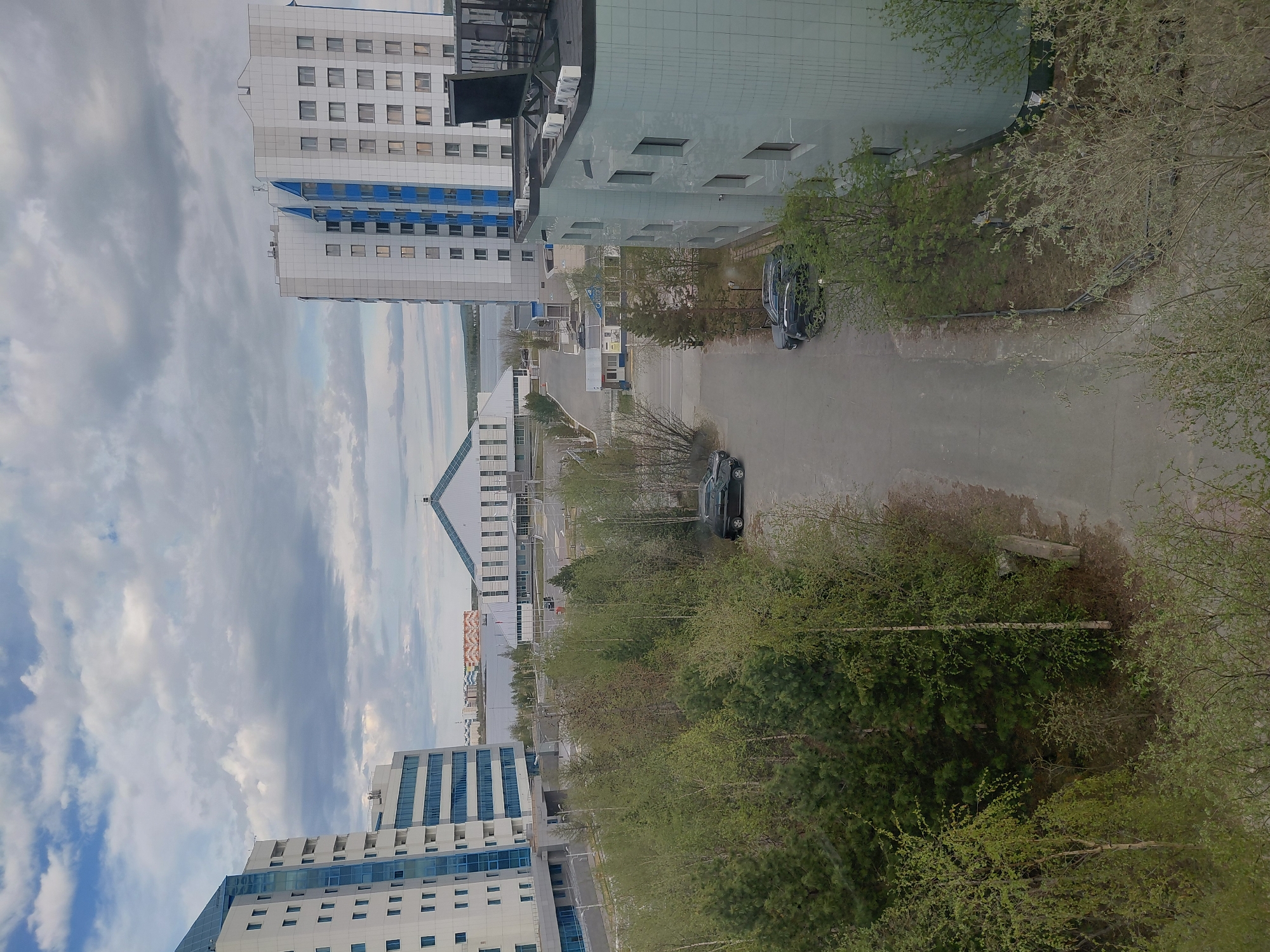 The image, captured from an elevated viewpoint, depicts a modern urban landscape in Nadym, Russia, under a cloudy daytime sky. The scene is dominated by several multi-story buildings featuring contemporary architecture.
In the foreground and mid-ground, distinct buildings showcase clean lines; some have prominent blue and white paneling, while others feature light grey or muted greenish-blue facades. A paved road or parking area winds through the scene, with at least two dark-colored sedans visibly parked on it. Extensive greenery, including trees with developing spring foliage and some evergreens, frames the road and buildings, particularly in the immediate foreground and mid-ground. One blue sign on the ground level shows partially visible, blurry text, which is unreadable.
In the background, beyond the immediate structures, a long, low-rise building with a distinct peaked roof can be seen. Further in the distance, a body of water, possibly a river or lake, stretches towards the horizon, bordered by fainter, more distant structures. The sky is predominantly overcast with varying shades of grey and white clouds. No people or specific activities are visible, presenting a calm, static urban environment that suggests planned development and modern infrastructure integrated with natural elements. The lighting indicates it is daytime, possibly late morning or afternoon.
