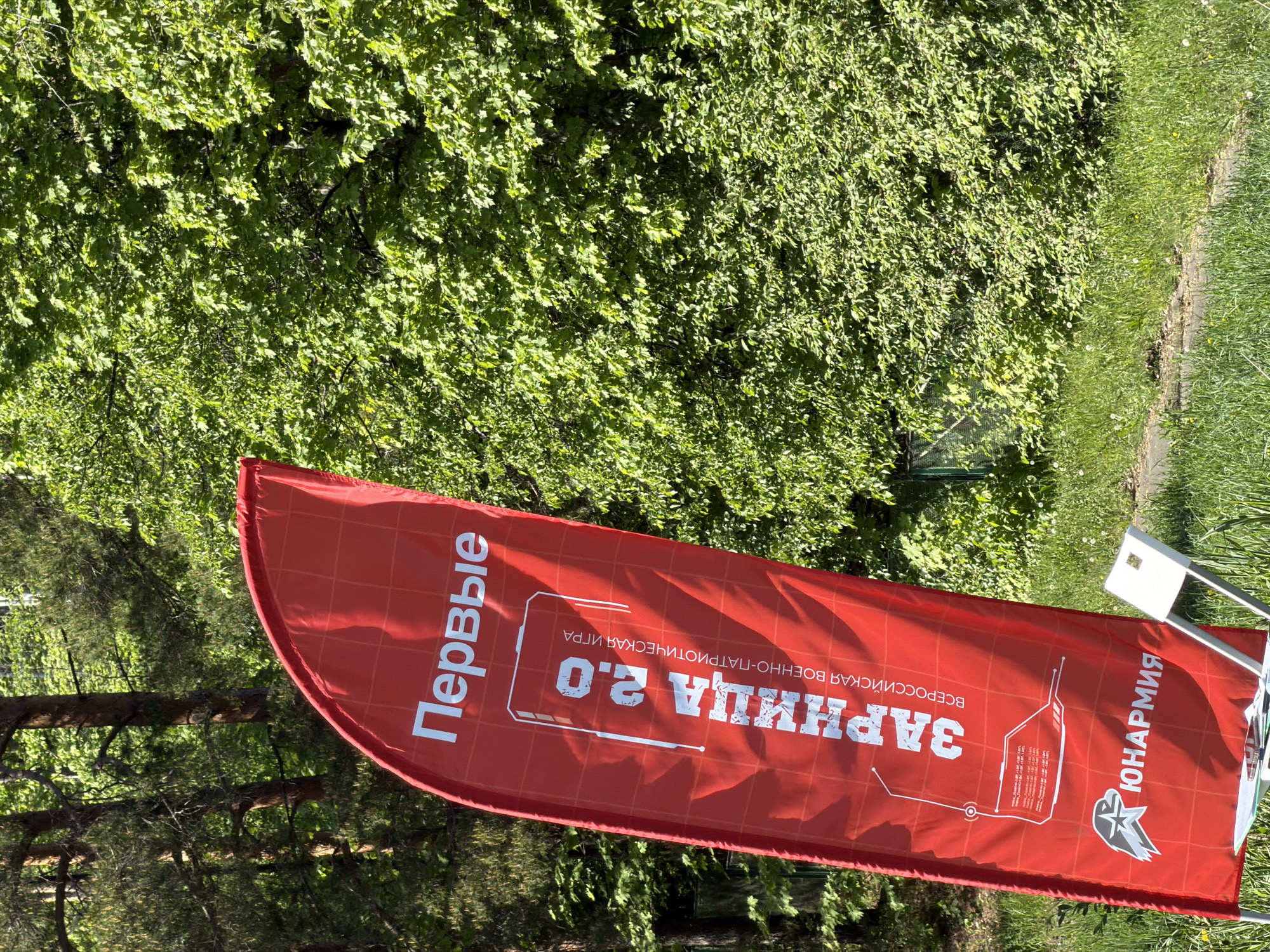 A red promotional flag is planted in a grassy area surrounded by trees, possibly Забегалово, Russia. The flag advertises "ЗАРНИЦА 2.0" (Zarnitsa 2.0), described as a "Всероссийская военно-патриотическая игра" (All-Russian military-patriotic game). The word "Первые" (First) is printed on the flag. The logo for "ЮНАРМИЯ" (Yunarmiya) is in the bottom right corner. The scene is outdoors in daylight, with sunlight filtering through the trees.