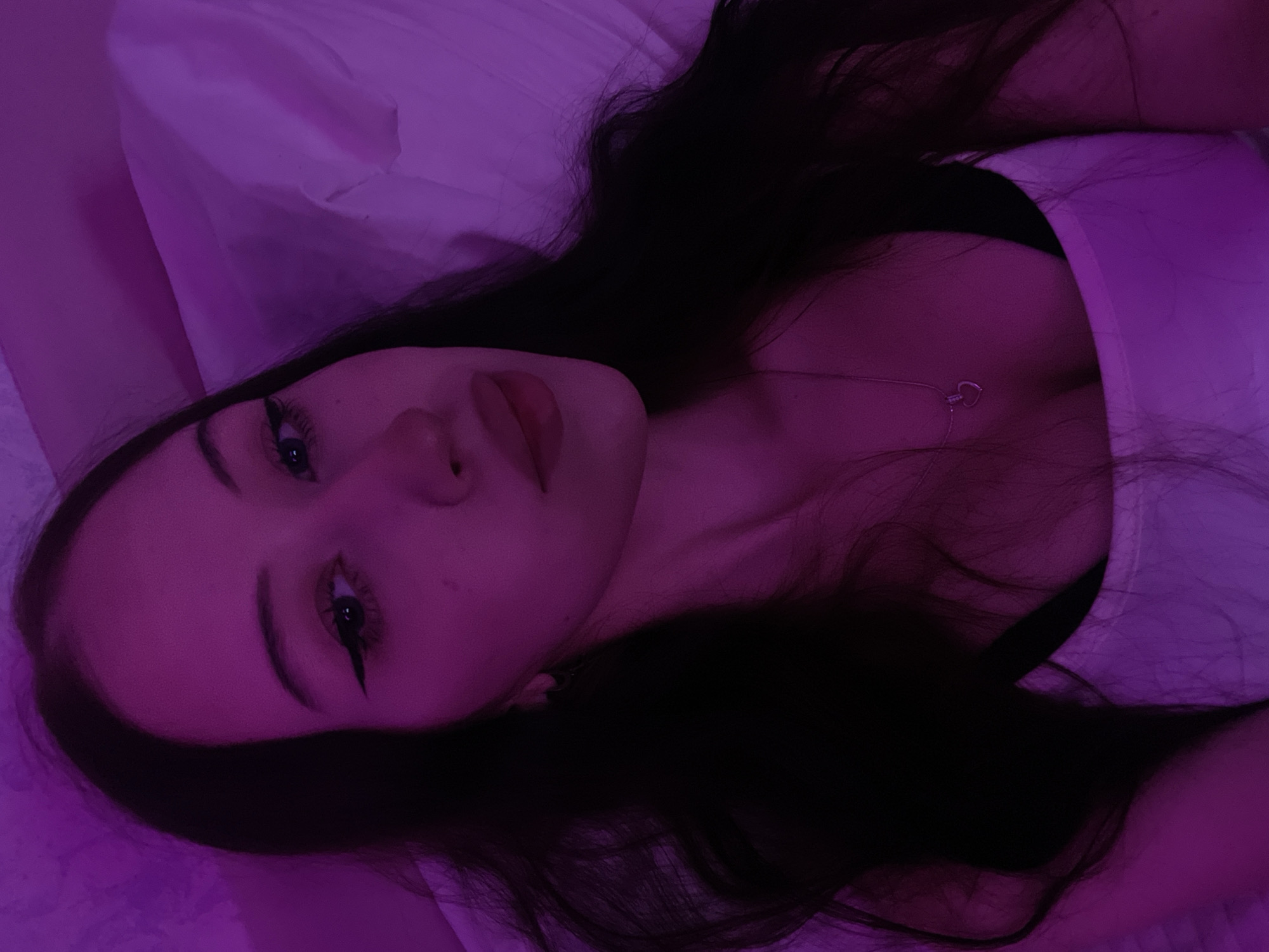 A young woman with long dark hair is lying in bed under purple lighting. She is wearing a white tank top and a silver necklace with a heart pendant. Her face is partially visible, and she has winged eyeliner on. The background consists of a rumpled white duvet and a purple light source. The scene appears to be indoors, likely a bedroom, and could be located in Votkinsk, Russia, based on the provided information. There are no other people or objects of significance in the image, and no specific actions are being taken by the subject.