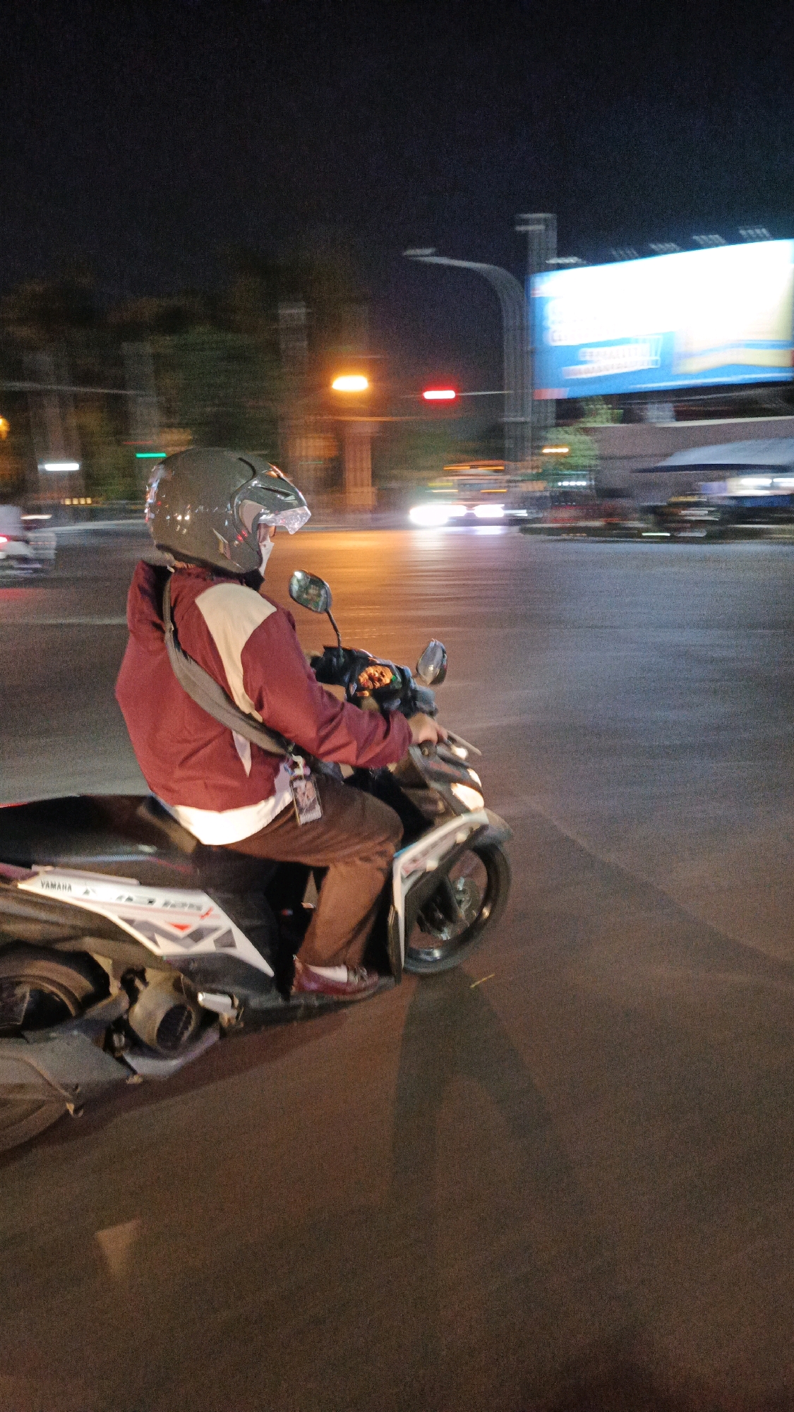 A person wearing a maroon jacket and brown pants rides a white Yamaha motorcycle at night on a street in Surakarta, Indonesia. The rider is wearing a helmet with the visor up and a white mask. The motorcycle has a visible "YAMAHA" logo on the side. The background shows a blurry cityscape with traffic lights, streetlights, and a large billboard advertising something on the right. The scene is illuminated by artificial lights, creating motion blur due to the movement of the motorcycle and surrounding traffic. The road surface is asphalt, and there are street markings visible.