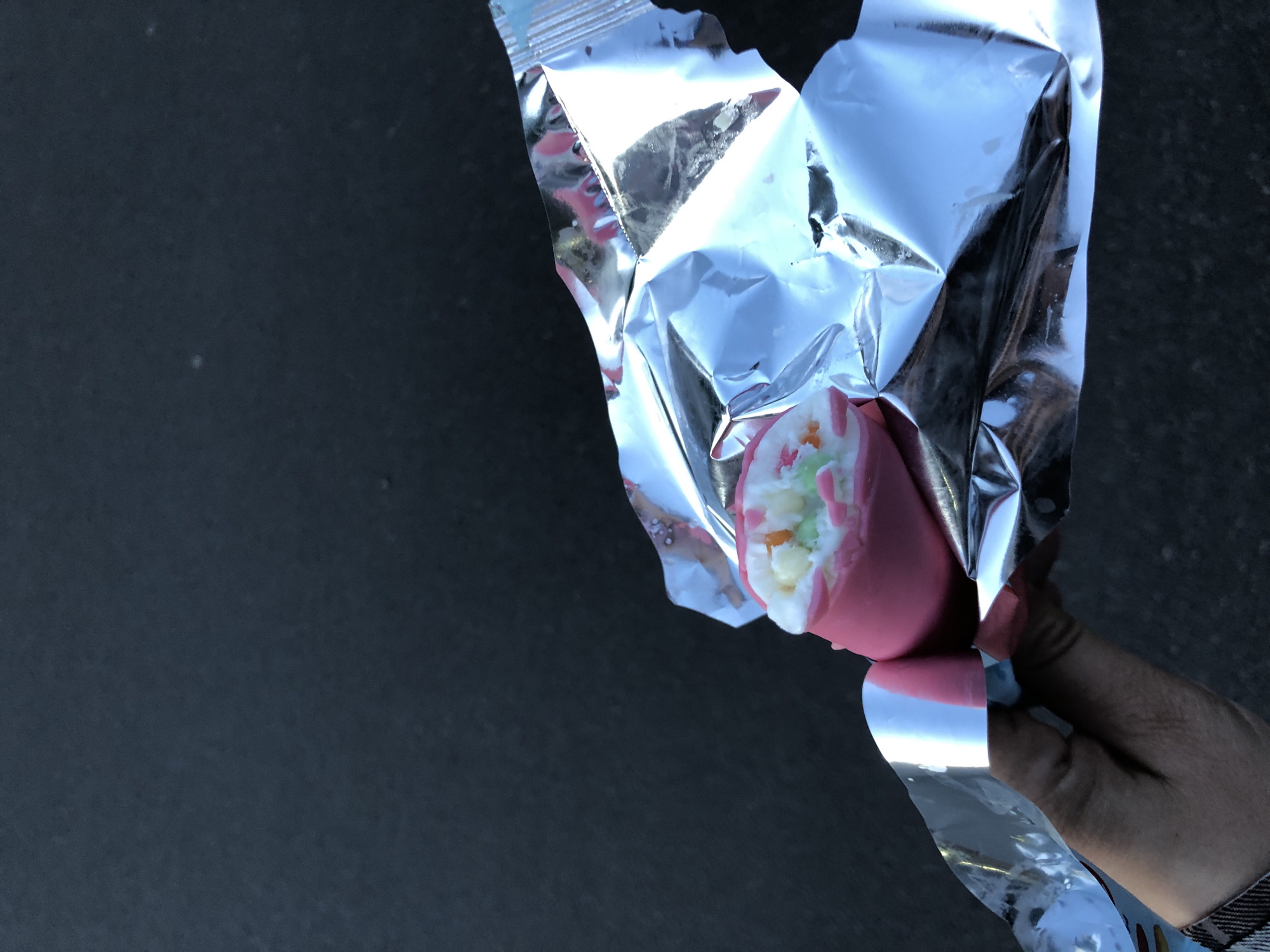 A close-up shot features a human hand holding a partially unwrapped confection against a dark, featureless background. The confection, which appears to be a sweet treat, has a smooth, pink outer layer. It is either bitten into or broken open, revealing a soft, white interior speckled with small, colorful pieces of green, orange, and white. This suggests a marshmallow-like consistency or a candy with various fillings. The treat is encased in a shiny, silver foil wrapper, which is crumpled from being opened. The hand, exhibiting a darker skin tone, is gripping the lower part of the wrapper and the treat. A small section of a patterned sleeve or clothing is visible around the wrist area. Bright reflections are visible on the metallic surface of the foil wrapper, indicating a light source. No text or specific branding is visible on the wrapper or the food item. The image captures the moment of unwrapping or consumption of the colorful dessert.