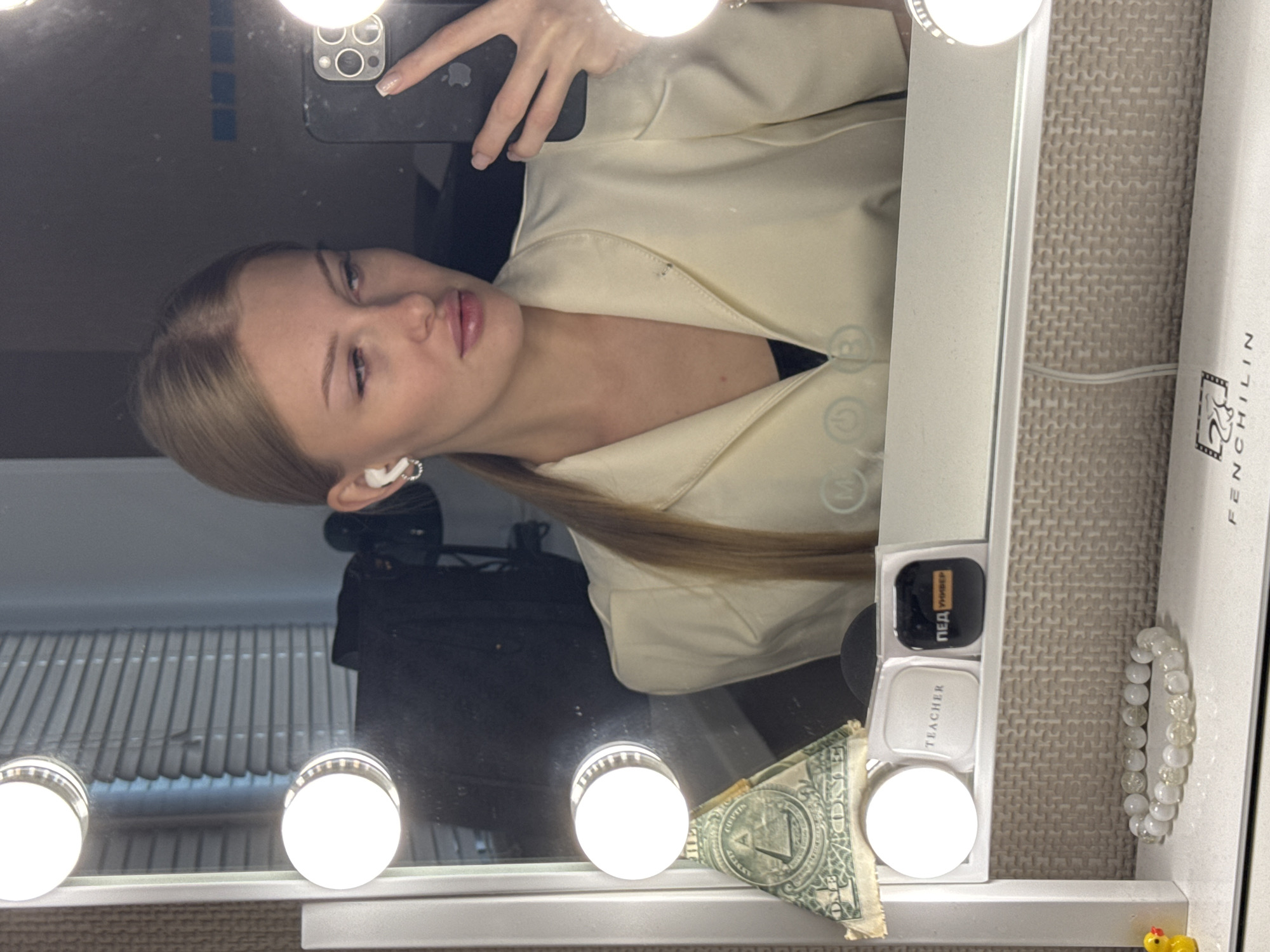 A young woman with blonde hair pulled back in a ponytail and wearing white earbuds takes a selfie in a mirror. She is wearing a light-colored blazer over a dark top. In the mirror's reflection, her phone is visible in her right hand. On a shelf in front of the mirror are several items, including a one-dollar bill folded into a pyramid shape, a small white compact labeled "TEACHER", and a black case with the word "PED" and "УНИВЕР" visible. Below the shelf, a beaded bracelet with white and clear beads is on display. The background appears to be an indoor setting with blinds covering a window. This scene is not identifiable as being in the city of Perm, Russia.