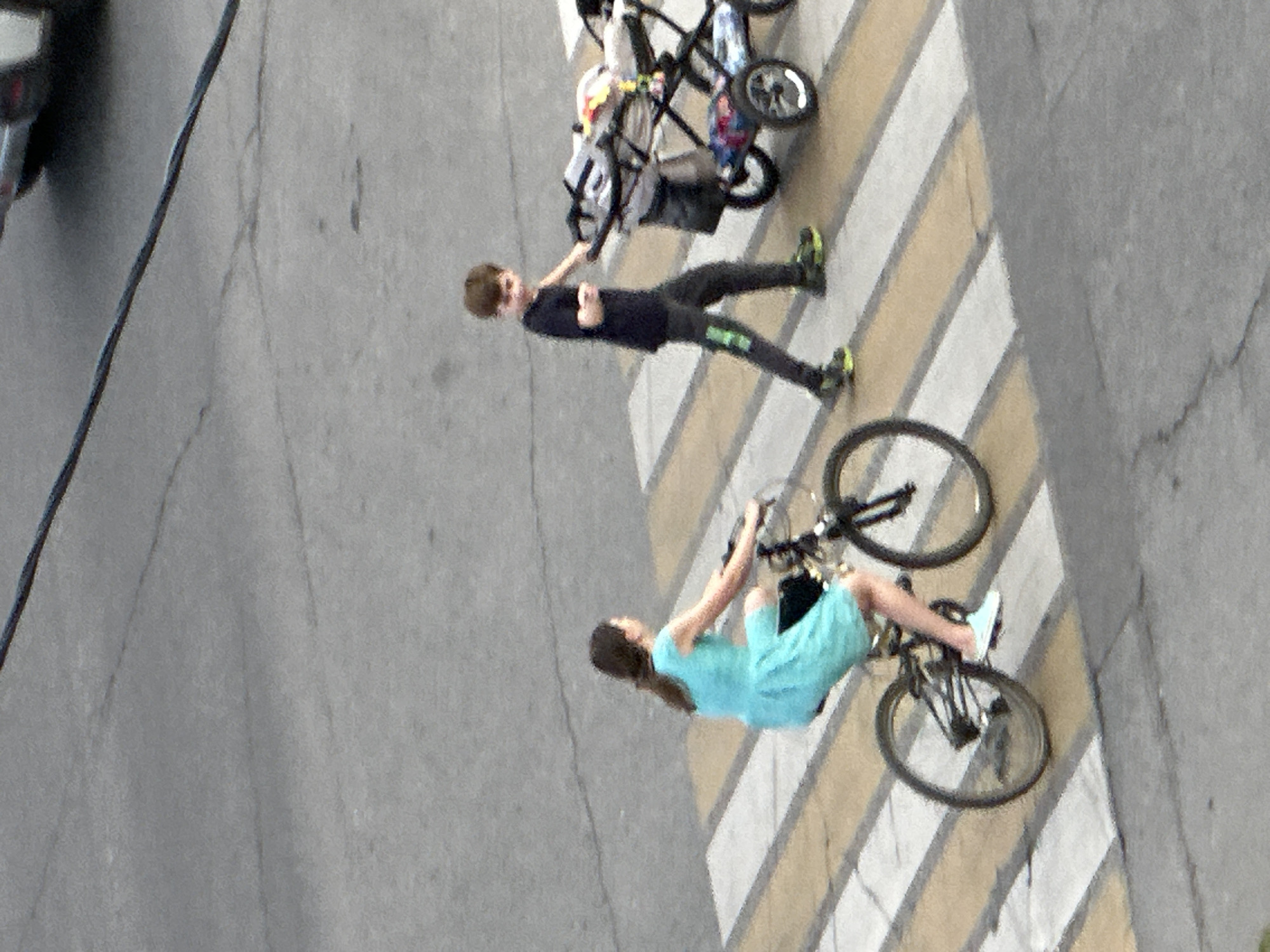 In Podolsk, Russia, at a pedestrian crossing, a young female is riding a bicycle. She is wearing a light blue dress and white sneakers. Her hair is dark brown and pulled back. To her right, a young male is walking and pushing a stroller. He is wearing a dark t-shirt, dark pants with a green stripe, and dark shoes with bright green accents. The stroller is grey and appears to contain some colorful items. The ground is asphalt with a visible crosswalk marked with white and yellow stripes. A dark wire is visible overhead. The lighting suggests it is daytime.
