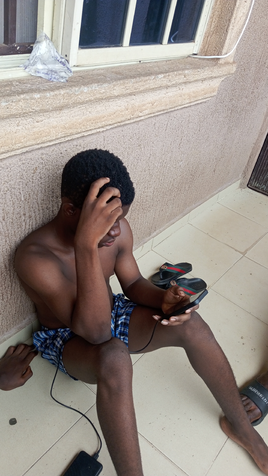 A young, shirtless man with short, dark curly hair sits on a light-colored tiled floor, leaning against a textured, light brown wall. He wears blue and white plaid shorts and holds a black smartphone in his hands, looking down at it intently. His right hand is raised, with fingers touching his forehead, suggesting contemplation or weariness. A black power bank is on the floor to his left, connected to the phone by a black charging cable.
Behind and to his right, a pair of black slides with a distinctive red and green stripe and "GUCCI" branding are visible on the floor. In the bottom right corner, another foot wearing a black slide with "SURBERRY" text is partially visible. In the bottom left, a hand or arm of another individual is seen on the floor.
Above the man's head, a light-framed window with dark glass or bars is present. On the window ledge, a clear plastic sachet of water with "BEST" and "Water 50cl" printed on it rests. A white cable also runs along the window sill to the right. The setting appears to be an indoor or covered outdoor area, illuminated by natural daylight.