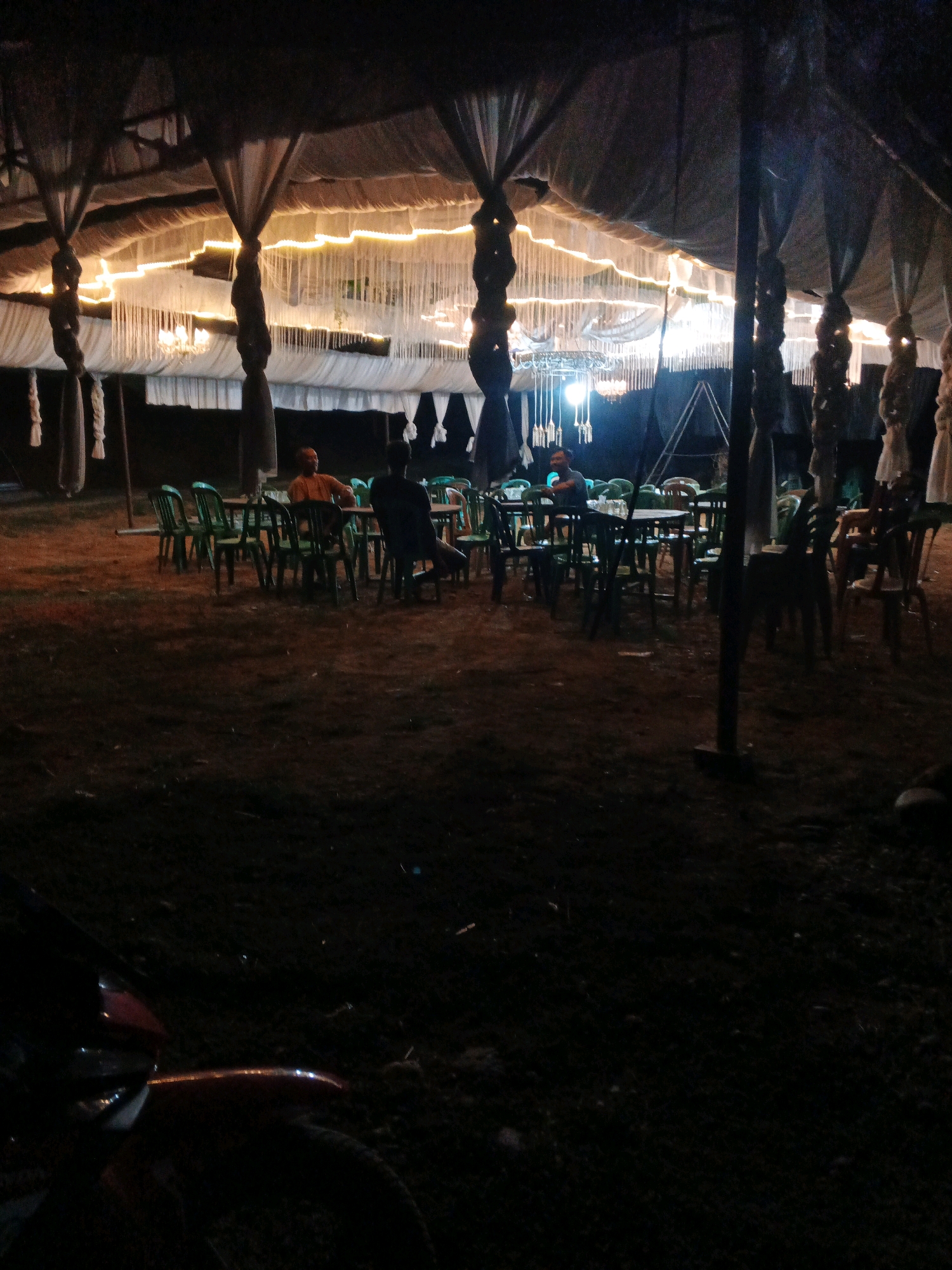 A nighttime outdoor scene under a tent structure with draped fabric and string lights. Several round tables and green plastic chairs are set up on the dirt ground. Four individuals are seated at two of the tables. One person in an orange shirt is sitting at a table on the left, facing away from the camera. To their right, another person is seated at the same table, facing the side. Further to the right, two more individuals are seated at a different table, both facing away from the camera. The tent is illuminated by warm white string lights and several chandeliers. The foreground is dark, with the back of a red and black motorcycle visible on the left. The overall impression is of a casual outdoor gathering or event. The location is identified as Brangan Mulya, Indonesia.