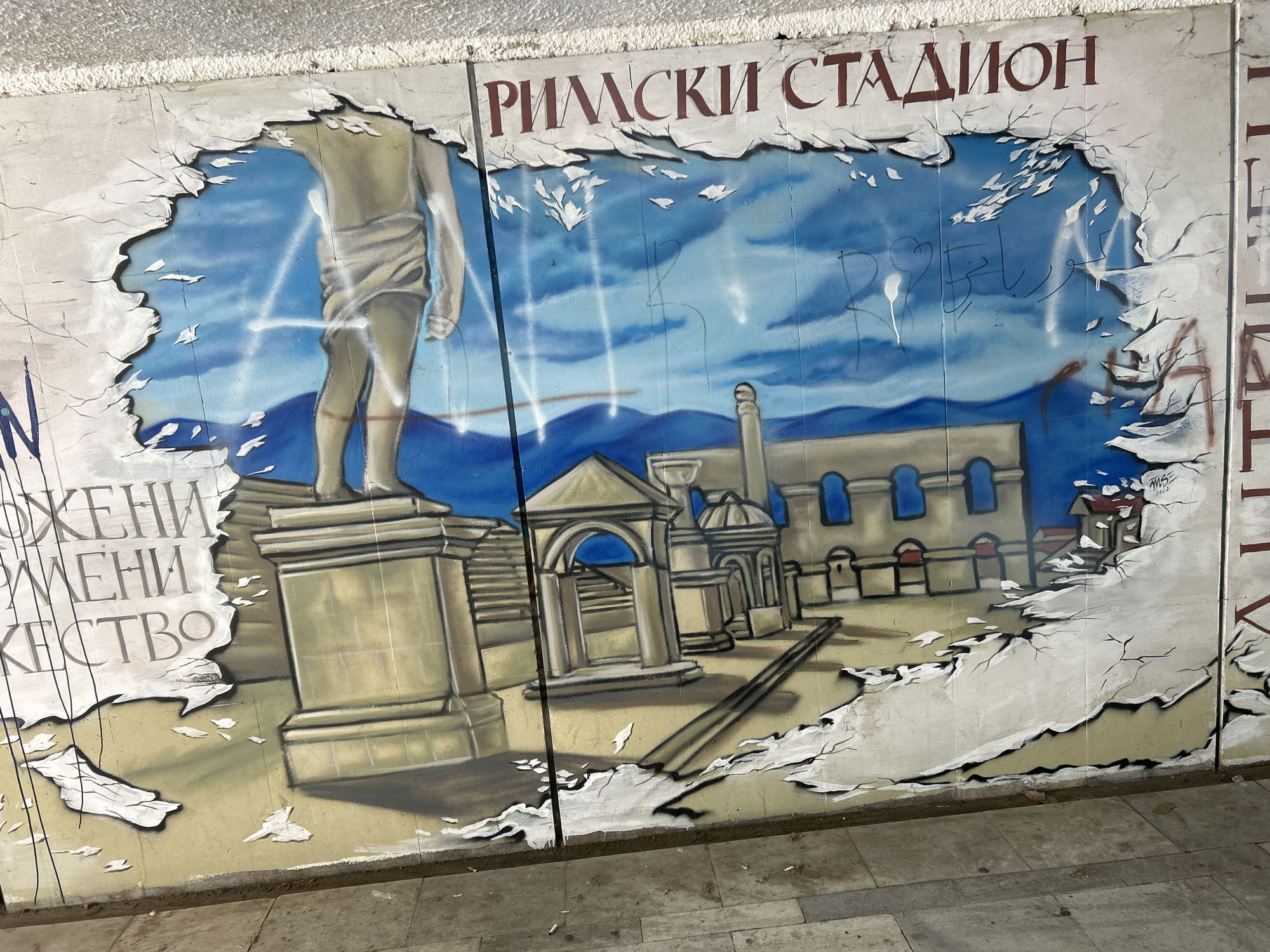 The image shows a mural in an underpass in Plovdiv, Bulgaria. The mural depicts the Roman Stadium, a historic site in the city. The name of the stadium, "Римски Стадион" (Roman Stadium) is written in Bulgarian above the mural. The mural is painted to appear as if it is breaking through the wall, with the edges of the painting depicted as torn. The scene is an exterior shot of the wall. The depiction includes a statue, architectural structures resembling ruins, and a background of blue sky and mountains. There is graffiti on the mural, indicating its age and exposure to the elements. The bottom of the mural is near the underpass floor.