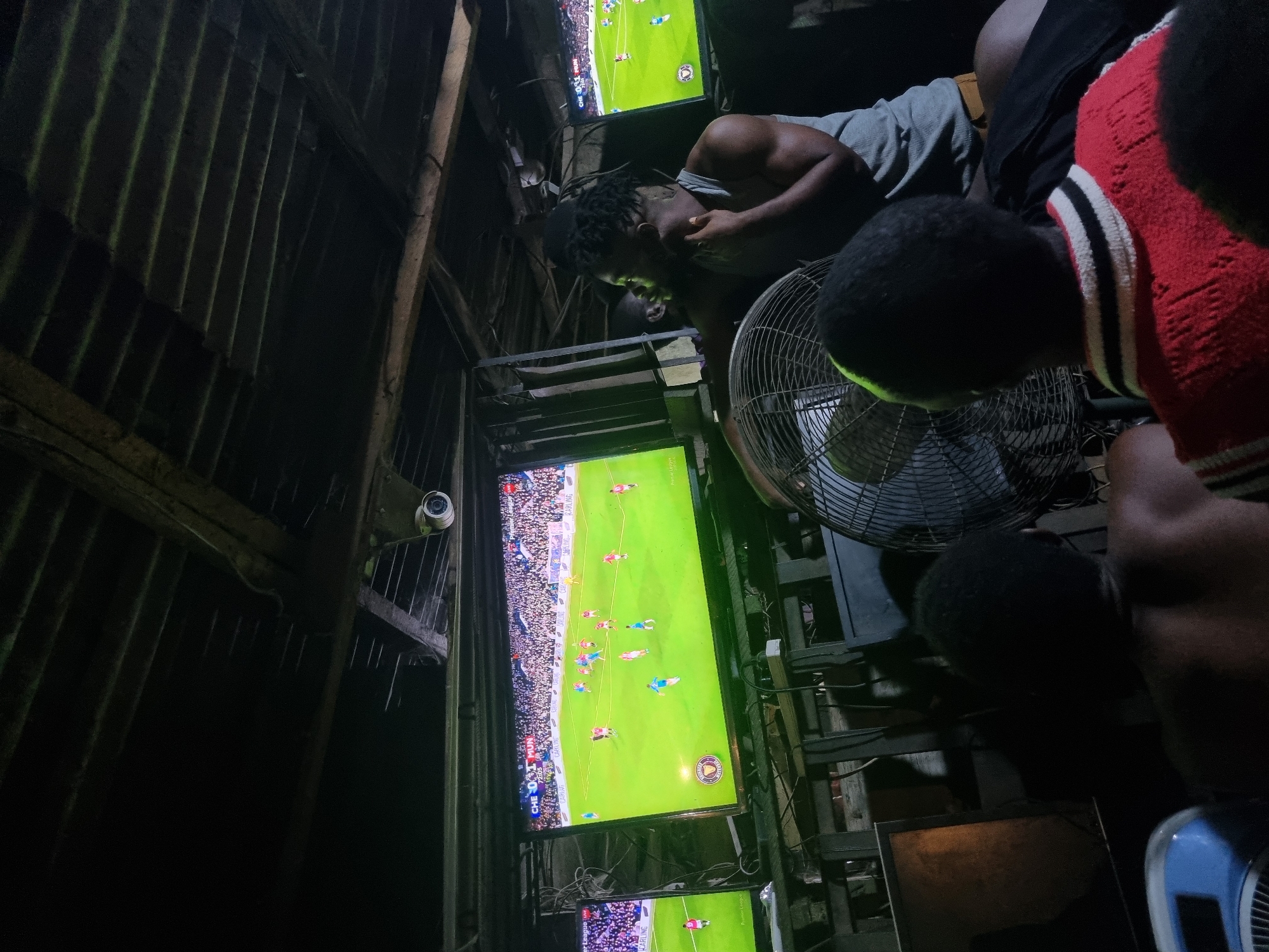 In a dimly lit, makeshift structure in Onitsha, Nigeria, a group of men are engrossed in watching a soccer game on multiple stacked television screens. The building appears to be constructed of rough-hewn wood and corrugated metal. A security camera is mounted on one of the beams. An oscillating fan is placed amongst them. The men are clad in casual clothing, including tank tops and a striped shirt. The mood seems intense, suggesting a critical moment in the game. Wires and electronic equipment clutter the background, highlighting the informal, functional nature of the space.