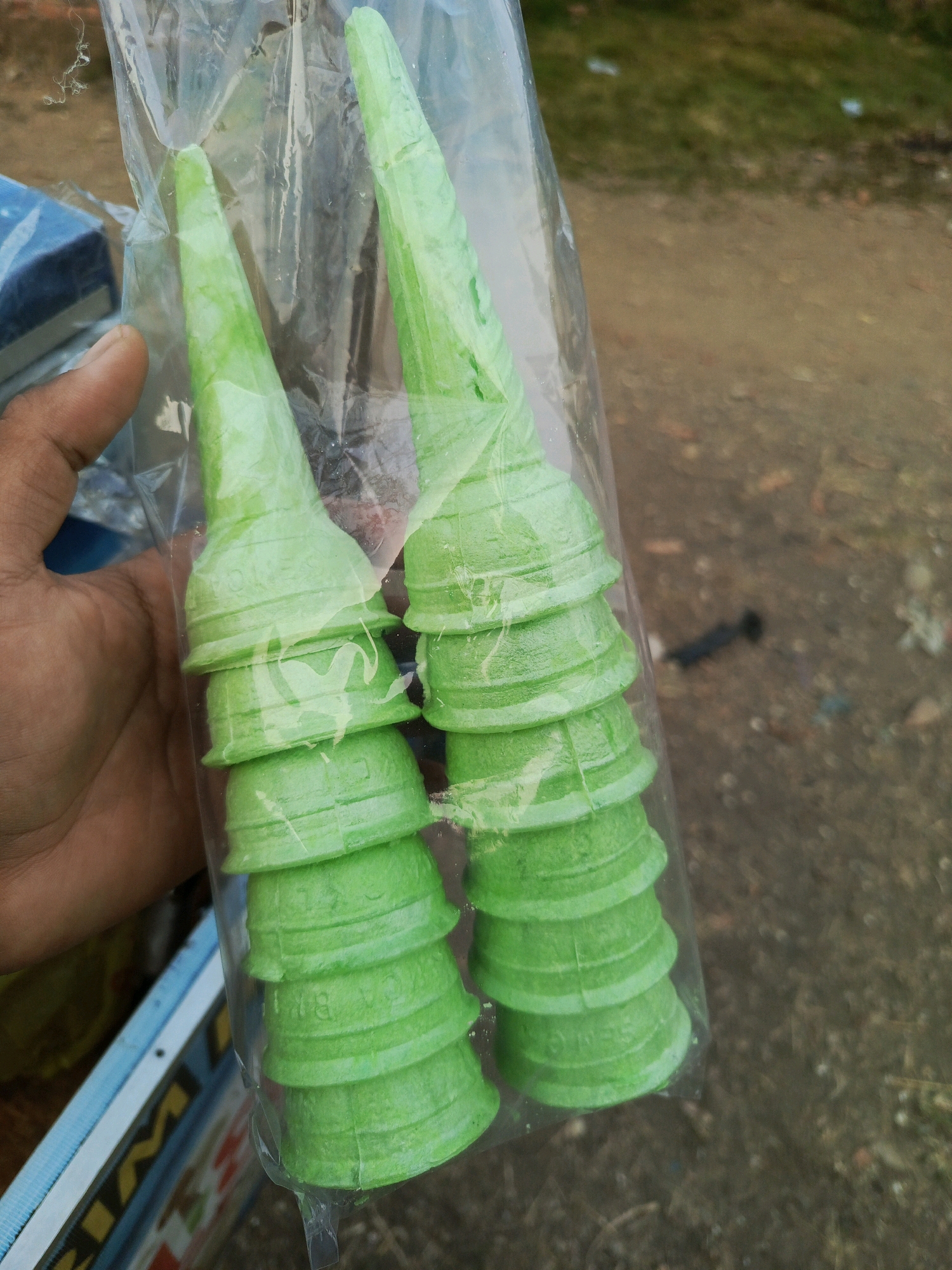 A hand is holding a clear plastic bag filled with stacked bright green ice cream cones. The cones are conical in shape with ribbed detailing around their base. The bag appears to be slightly crinkled, and a portion of a blue and white striped object is visible in the lower left corner, partially obscured by the hand and the bag. The background is blurry, suggesting an outdoor environment with brown dirt or gravel and patches of green grass. There are no visible people or specific landmarks that would indicate the location of Megang Sakti II, Indonesia.