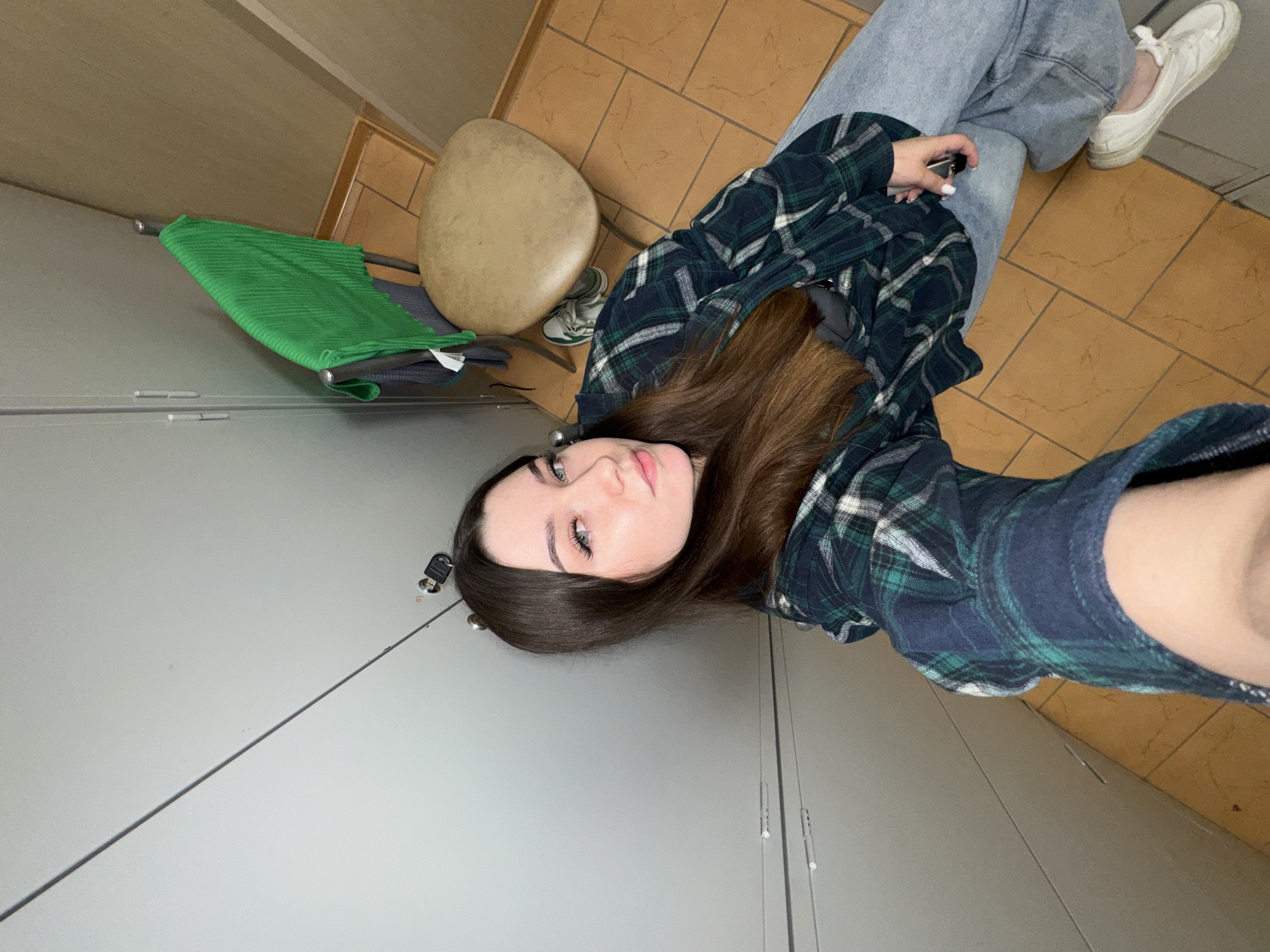 A young woman with long brown hair is taking a selfie in a locker room. She is wearing a green and blue plaid shirt and light blue jeans. She is holding a key in her right hand. In the background, there are grey lockers, a chair with a green towel draped over it, and a tan-tiled floor. The scene appears to be in a public facility such as a gym or a school. The location is identified as Zarechnaya (Gamovskoe S/P), Russia.