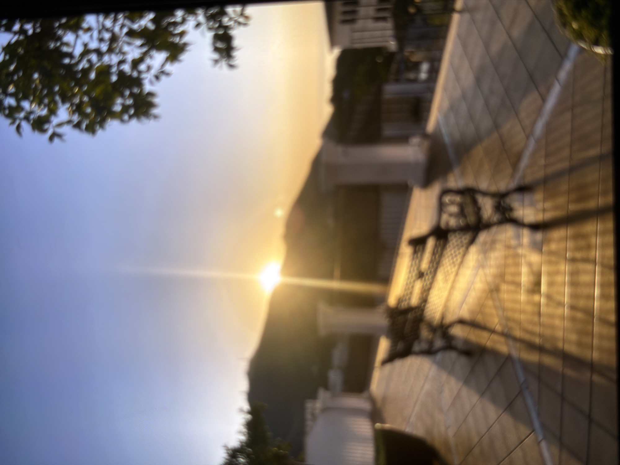 This is a photograph of a sunset viewed from a tiled balcony. The sun is a bright, diffused orb low in the sky, casting a warm, golden light. The sky transitions from a pale blue to a bright yellow near the horizon. In the foreground, a dark, wrought-iron bench is visible on the tiled surface. Shadows from the bench and possibly foliage stretch across the tiles. To the left, out-of-focus green foliage hangs into the frame. In the background, a blurred silhouette of a hill or mountain is discernible, with some architectural elements suggesting buildings. The overall mood is peaceful and serene, evoking the end of a day. The time of day is clearly sunset. The weather appears clear and calm.