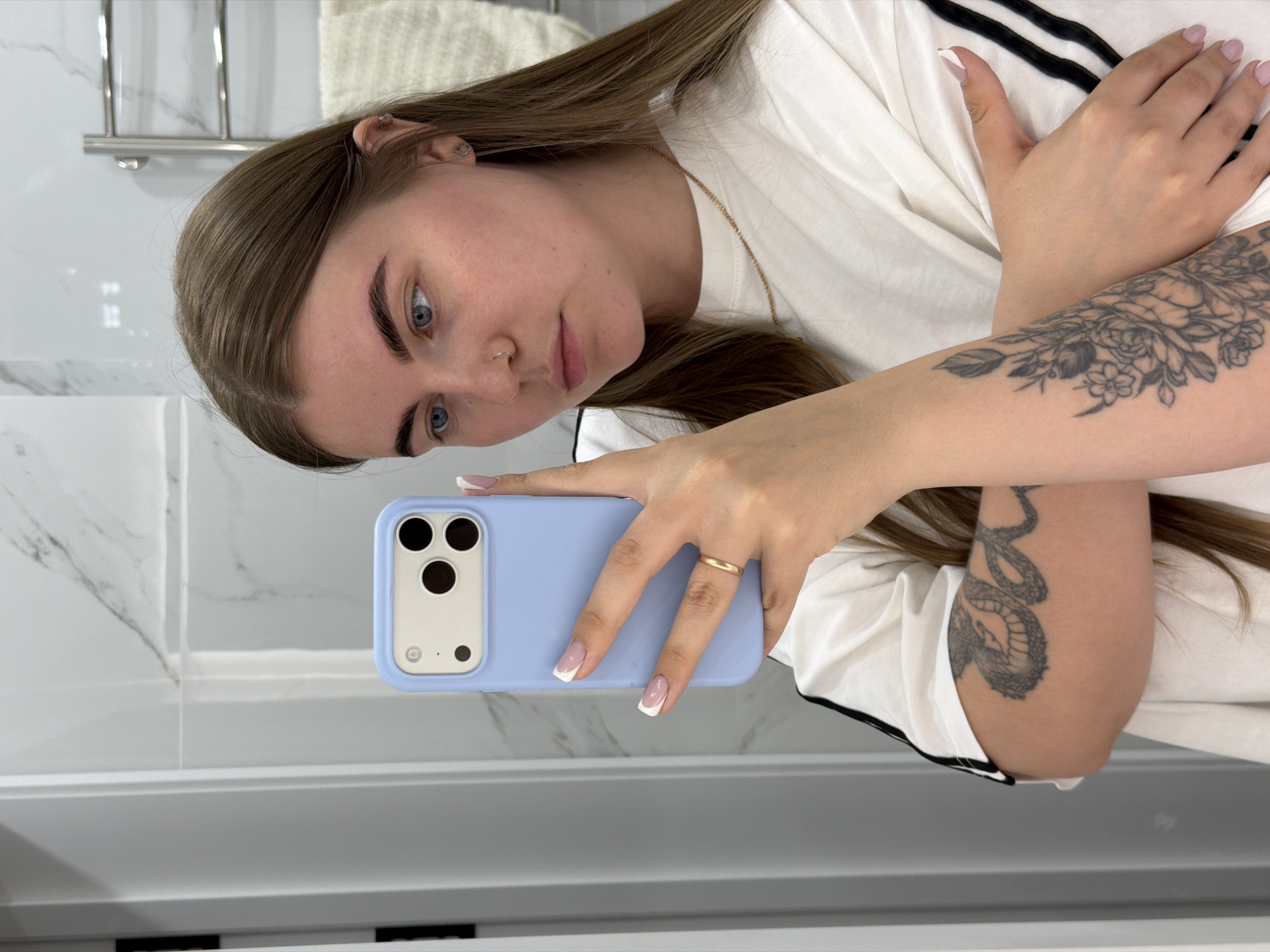 A young woman with long brown hair is taking a selfie in what appears to be a bathroom. She is wearing a white shirt with black stripes on the sleeves and holding a blue smartphone. She has tattoos on her arms, one featuring floral designs and another depicting a snake. A gold ring is on her ring finger. The background shows a white wall, possibly with a marble-like texture, a towel rack, and part of a white sink or countertop. There is no information to indicate that this image was taken in Chaikovsky, Russia.
