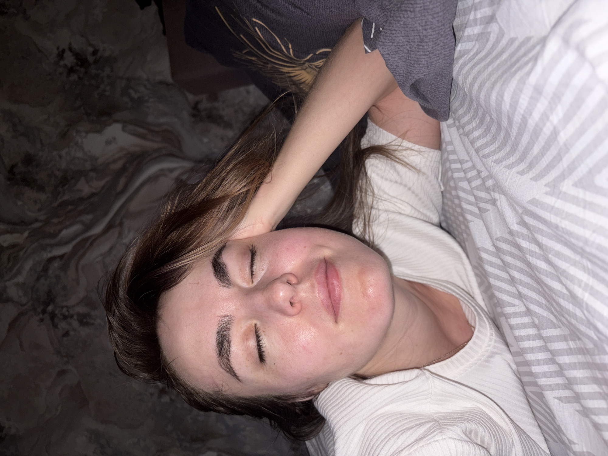 A woman with light skin and closed eyes is lying down on a bed. She is wearing a white, ribbed shirt and a thin silver necklace. Her brown hair is parted on one side, with some strands touching her forehead. Her left arm is raised and bent, with her hand near her head. A gray sleeve is visible near her arm. The bed has gray and white striped sheets. A dark gray pillow with a golden design is visible in the background. The background floor has a dark, marbled pattern. There is no specific information to suggest the location is Izhevsk, Russia.