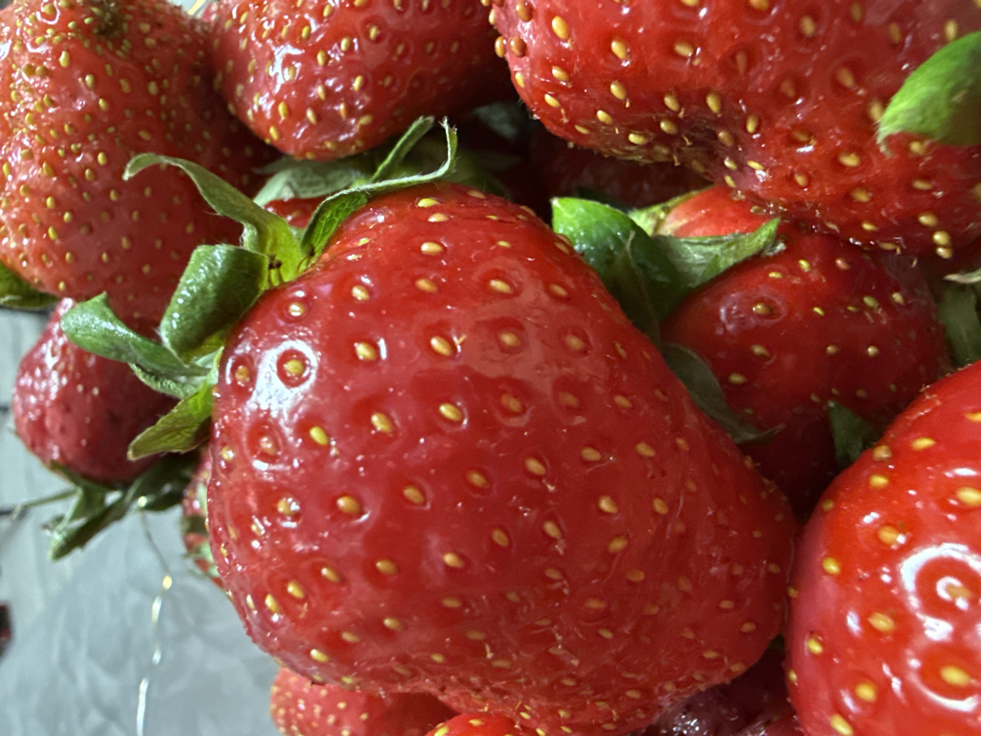 The image shows a close-up of a pile of red strawberries with green leaves. The strawberries are ripe and have numerous small, yellow seeds. The fruit appears fresh and glossy. The location is unknown, and there is no information in the image to confirm it is in Chaikovsky, Russia. No people, subjects, objects, actions, or interactions are depicted other than the strawberries themselves. The environment is a soft, neutral-toned surface upon which the strawberries rest.