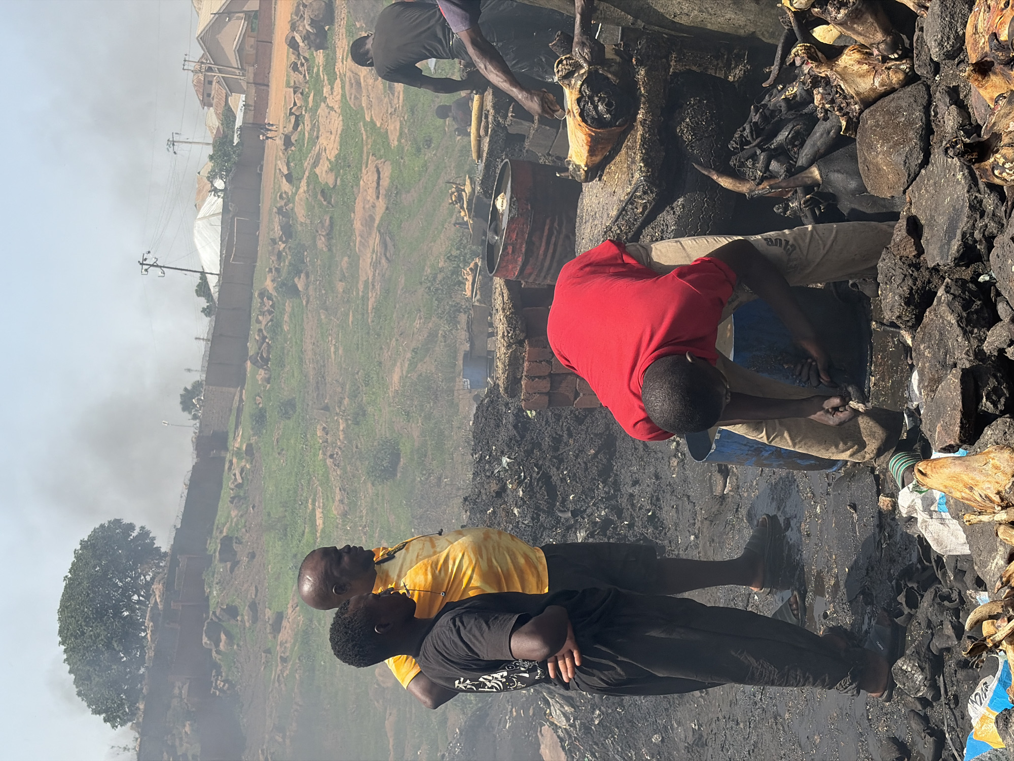 In Jos, Nigeria, the image depicts a scene with several individuals working amidst the charred remains of what appears to be animal parts and a smoky environment. At least four people are visible, one in a red shirt leaning over the debris, another in a yellow shirt standing with a younger person in a black shirt. A fourth person can be seen further back also working at the fire. The ground is covered in ash and remnants of burnt material, suggesting a recent fire or ongoing burning process. In the background, there is a landscape with vegetation and some buildings, indicating an urban or semi-urban setting.