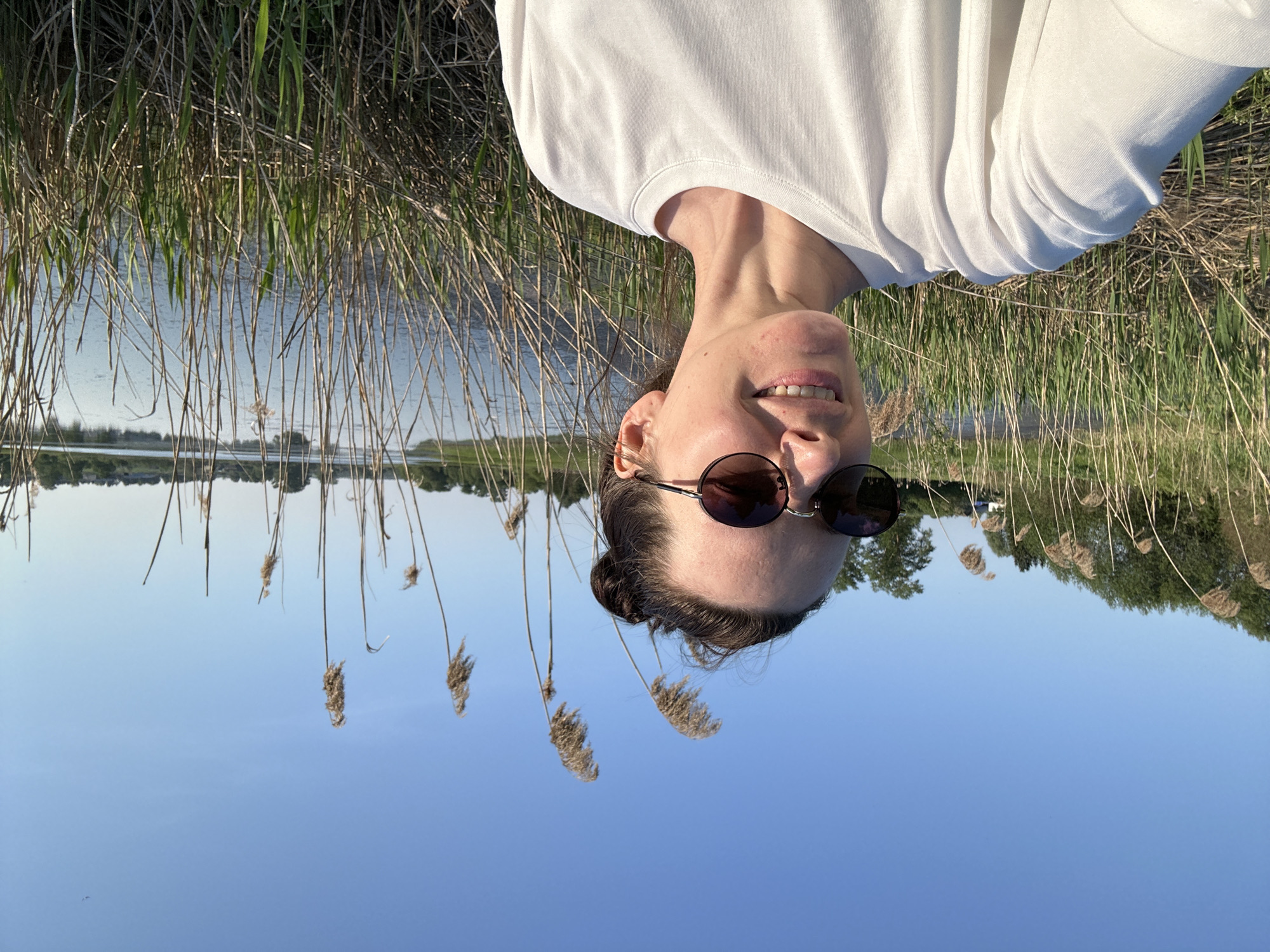 A photo shows a woman wearing sunglasses and a white shirt. The photo appears to be taken in Альняш, Russia near a lake and reeds. The background reflects off of the water. The photo has been rotated.