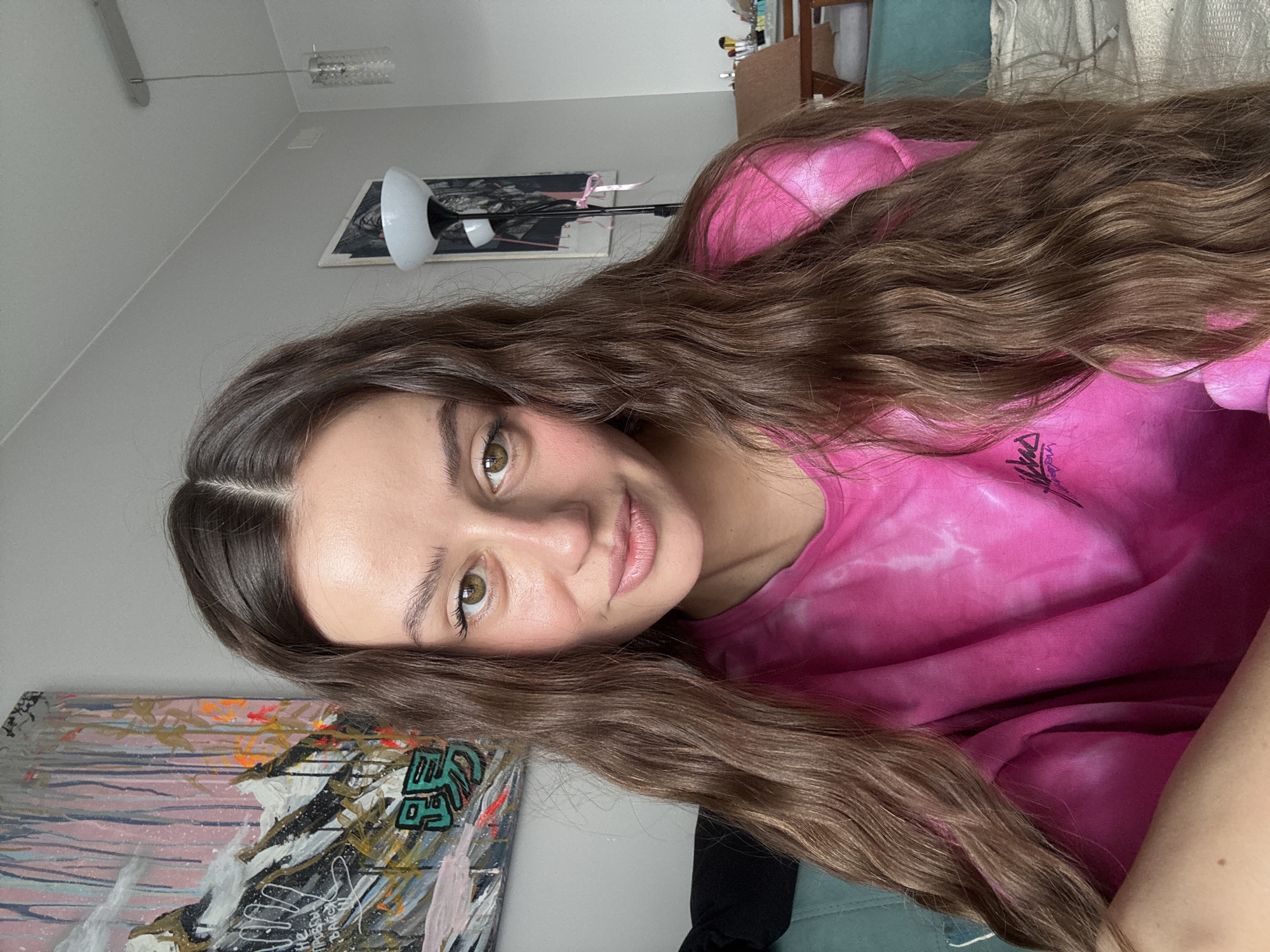 A woman with long, wavy brown hair is facing the camera. She has fair skin, brown eyes, and is wearing a pink tie-dye shirt. The background includes a painting with various colors, a floor lamp, a picture on the wall, a ceiling fan, and furniture. The location cannot be determined from the image.