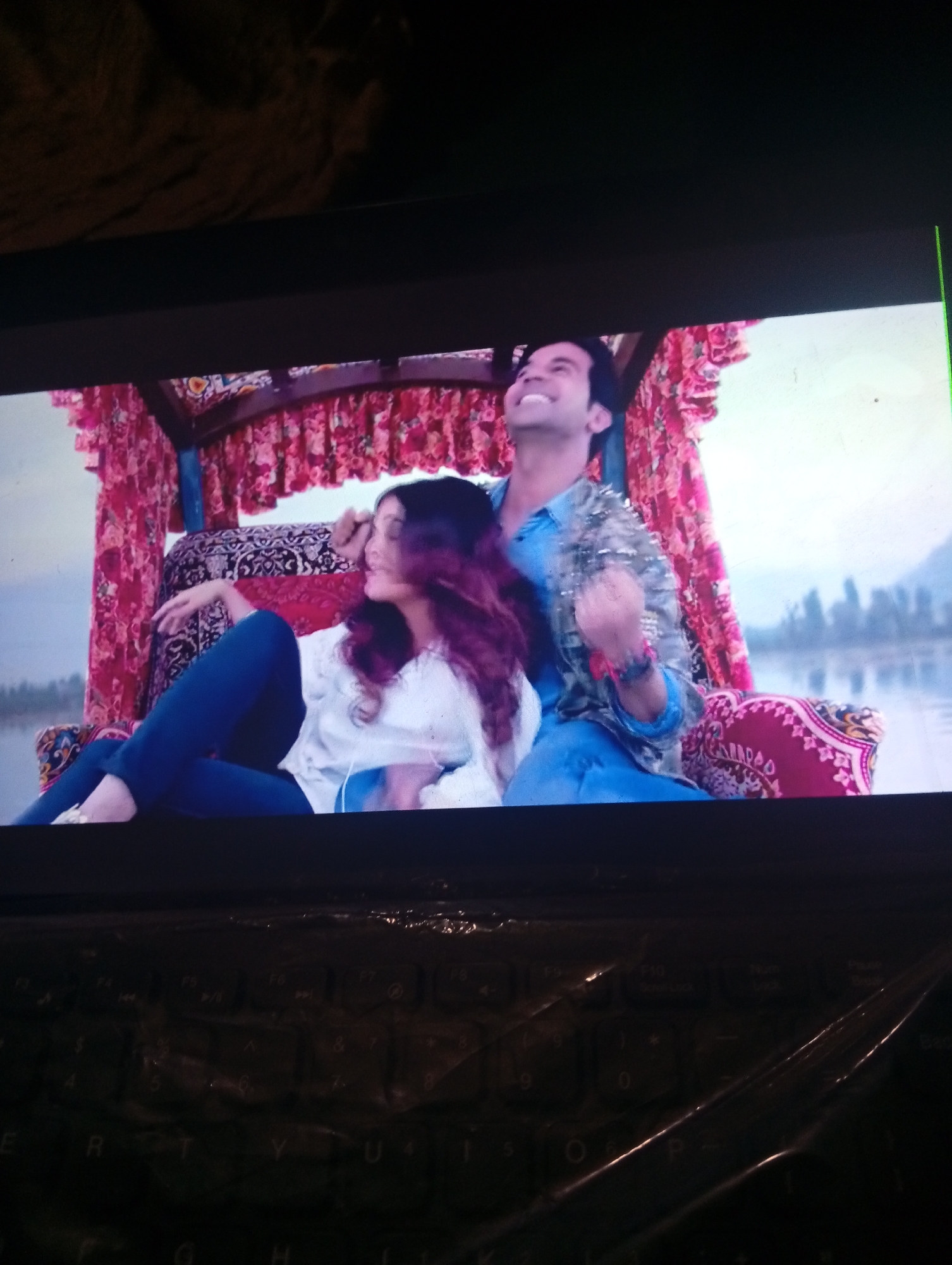 A man and a woman are seated in a decorated boat. The woman, on the left, has long, purple-tinted hair and is wearing a white top and dark blue pants. She is leaning back, with her left arm resting behind her head. The man, on the right, is wearing a blue shirt and has a beaded or sequined jacket over it. He is looking upwards and smiling. The boat has a red and pink floral canopy and ornate cushions. In the background, there is a body of water with trees and hills visible on the horizon. The scene appears to be a still from a movie or television show. The image is a photograph of a screen displaying this scene, with a keyboard visible at the bottom. The location of Azume, Nigeria is not discernible from the visual information in the image.