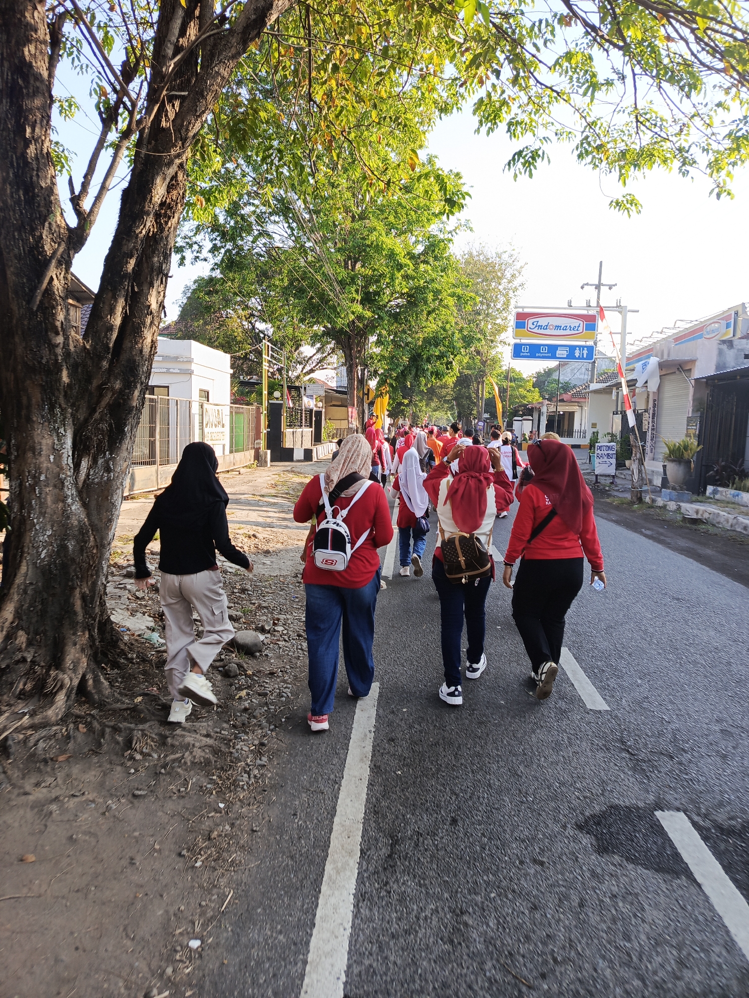 Four individuals, viewed from behind, walk along a paved road in Patokan, Indonesia. The person furthest to the left wears a black headscarf, a black long-sleeved shirt, and beige cargo pants with white sneakers. To their right, a person in a red shirt and blue jeans wears a white backpack with a black logo and a light-colored headscarf. Next to them, a person in a red long-sleeved shirt and black pants wears a red headscarf and carries a water bottle. The fourth person, also in a red long-sleeved shirt and black pants with a red headscarf, has a brown backpack with a gold logo. In the background, a cluster of people in red attire walks further down the road. Trees with green foliage line the left side of the road, casting shadows. Buildings, including one with an "Indomaret" sign and another with a sign indicating "POTONG RAMBUT," are visible on the right side. The road is marked with a white line and has some debris scattered along its edge. The sky is bright and appears to be clear.