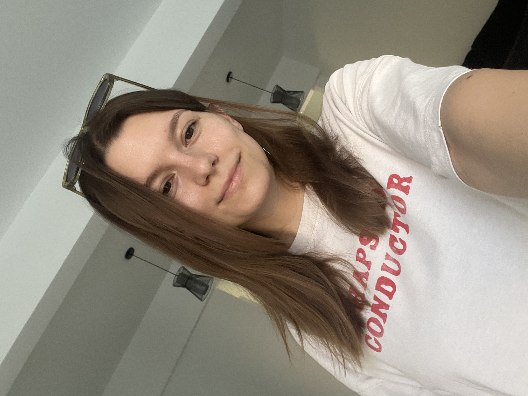 A woman with shoulder-length brown hair and sunglasses perched on her head is captured in a close-up, selfie-style shot. She is wearing a white t-shirt with red lettering that reads "NAPS CONDUCTOR." She has light skin and appears to be indoors, with ceiling lights and a muted background visible. The image seems to be taken in a residential or indoor setting, possibly in Limassol, Cyprus.