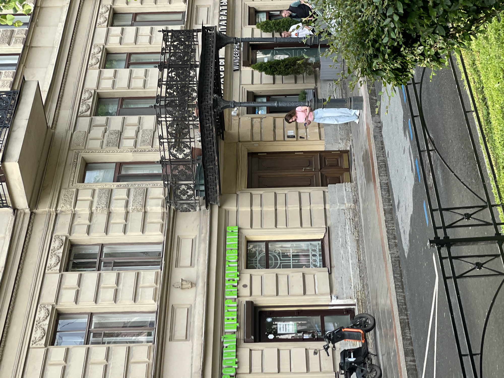 The image captures an urban street scene in Saint Petersburg, Russia, featuring a grand, light-colored multi-story building facade on the left. The building displays classical architectural elements, including decorative relief patterns around its numerous windows and an elaborate black wrought-iron balcony extending from an upper floor.
On the ground level, a young woman with light brown hair, dressed in a pink top and light blue jeans, stands on the paved sidewalk, looking down at her phone. Nearby, a black lamppost stands tall. Further along the building, towards the right, two men are visible near a dark wooden entrance door, partially obscured by a green bush. One man wears a white t-shirt, the other a dark one.
A black motor scooter or moped is parked on the sidewalk in the lower right portion of the frame. The street and parts of the sidewalk appear damp, indicating recent rainfall. A black metal fence separates the paved area from a lush patch of green grass and foliage on the far right.
Several signs are visible on the building. Above the main entrance, "WUNDERPHC" is displayed, alongside the partially visible Russian word "АПАРАТЫ" (Apparatus), suggesting an electronics or repair shop. A prominent vertical green sign on the lower part of the facade reads "РОДНОЙ ЦЕНТР РЕМОНТОВАН..." (Native Repair Center...). The scene unfolds during daytime, characterized by soft, possibly overcast, lighting.