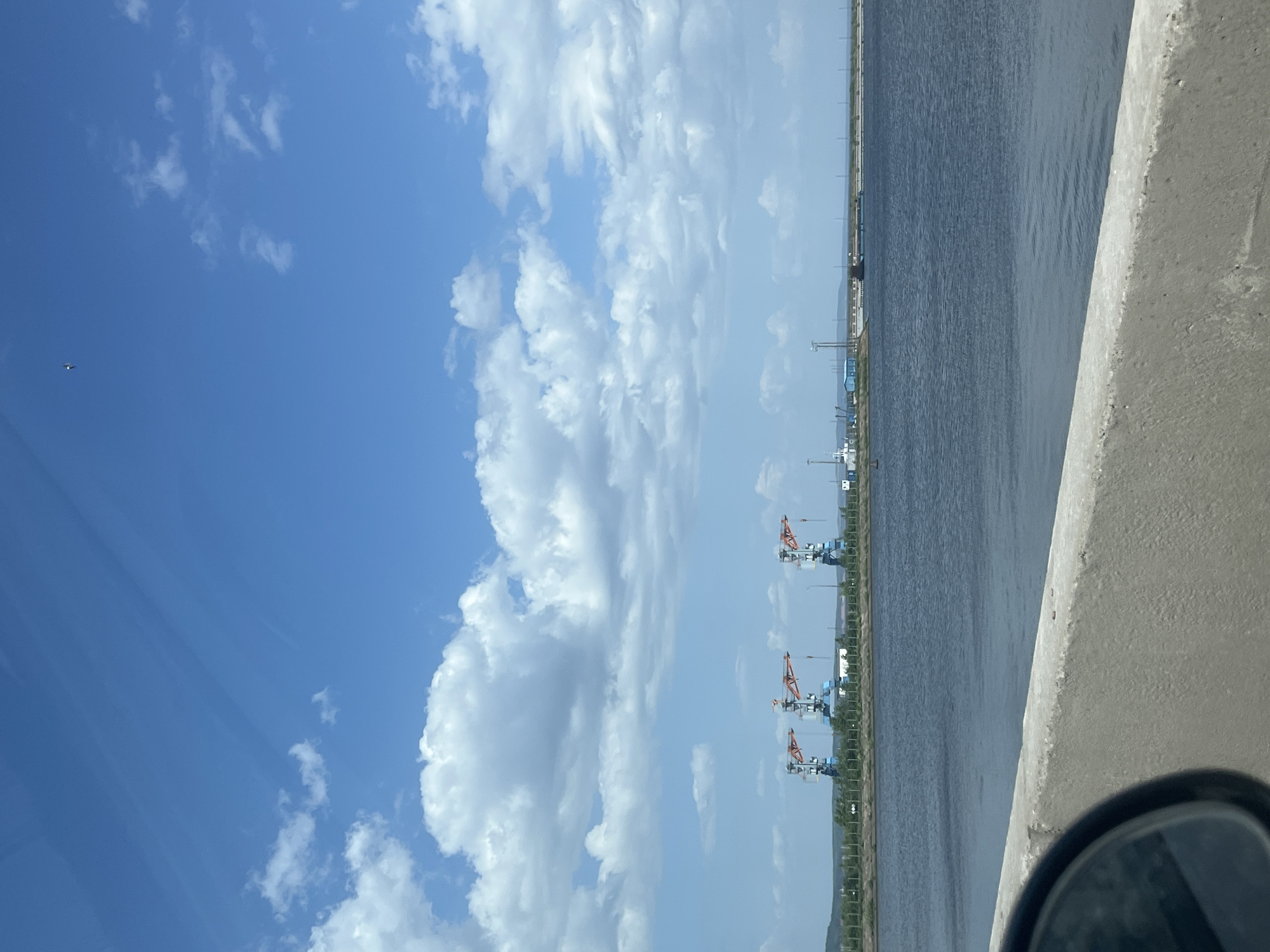 This image shows a view over the Kama River in Chaikovsky, Russia. The perspective is likely taken from a high point, perhaps a bridge or elevated roadway. The sky is mostly blue with scattered cumulus clouds. The water surface is dark and calm, with a faint ripple pattern. Along the far bank of the river, there are a series of cranes. A gray concrete barrier is in the right foreground. The corner of a car side mirror is visible.