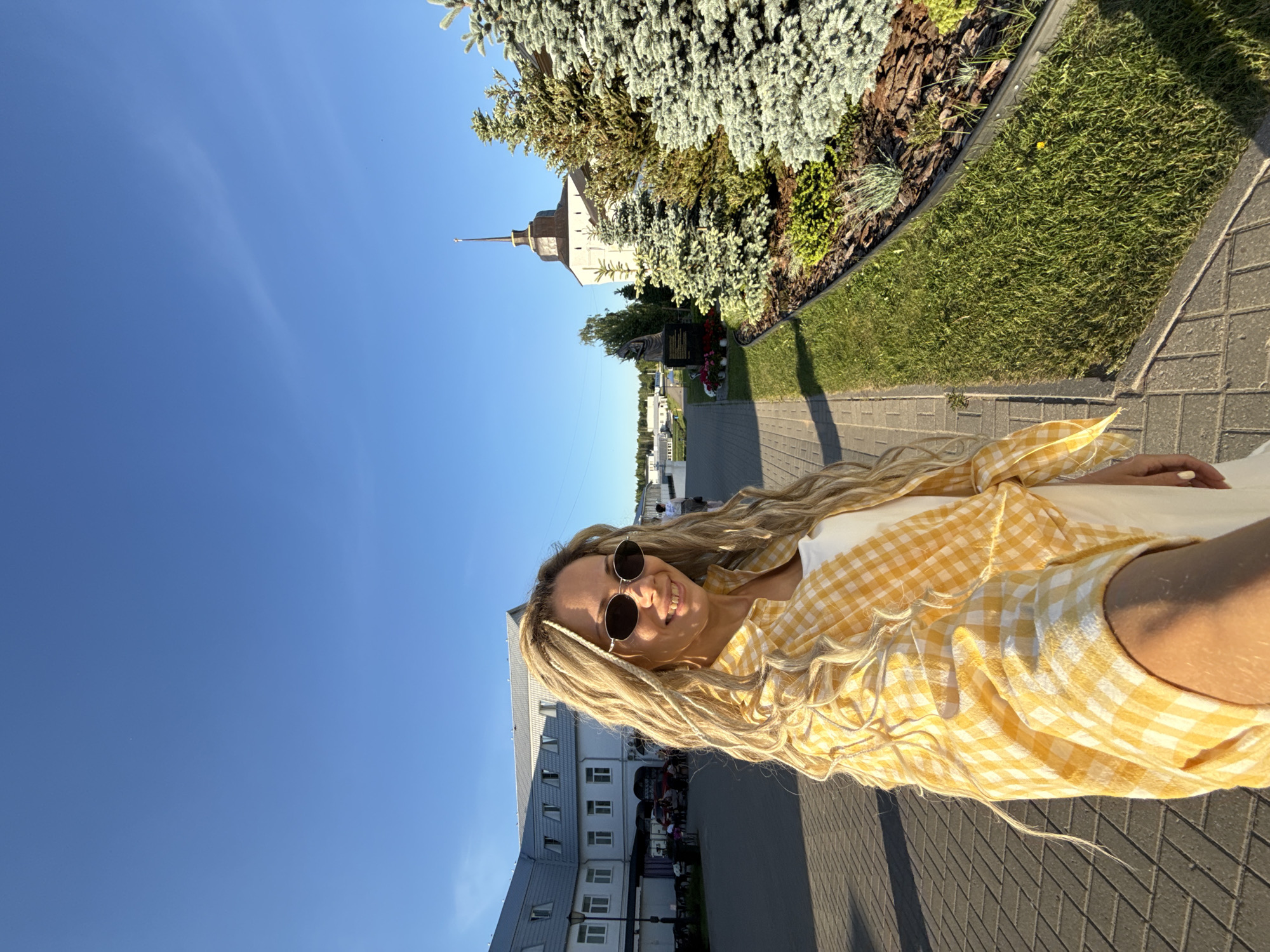 A smiling blonde woman with dreadlocks and sunglasses takes a selfie on a paved walkway in Rusanovo, Russia, under a clear blue sky. She wears a white top and a yellow and white checkered shirt. To her left, a large white building with a gray roof features a cafe area with outdoor seating. To her right, a manicured green lawn abuts a landscaped bed with various evergreen trees and ground cover. Further right, a bronze statue stands next to a white building with a distinctive brown, domed roof and a tall, pointed spire. The scene is illuminated by bright sunlight, casting shadows on the ground.