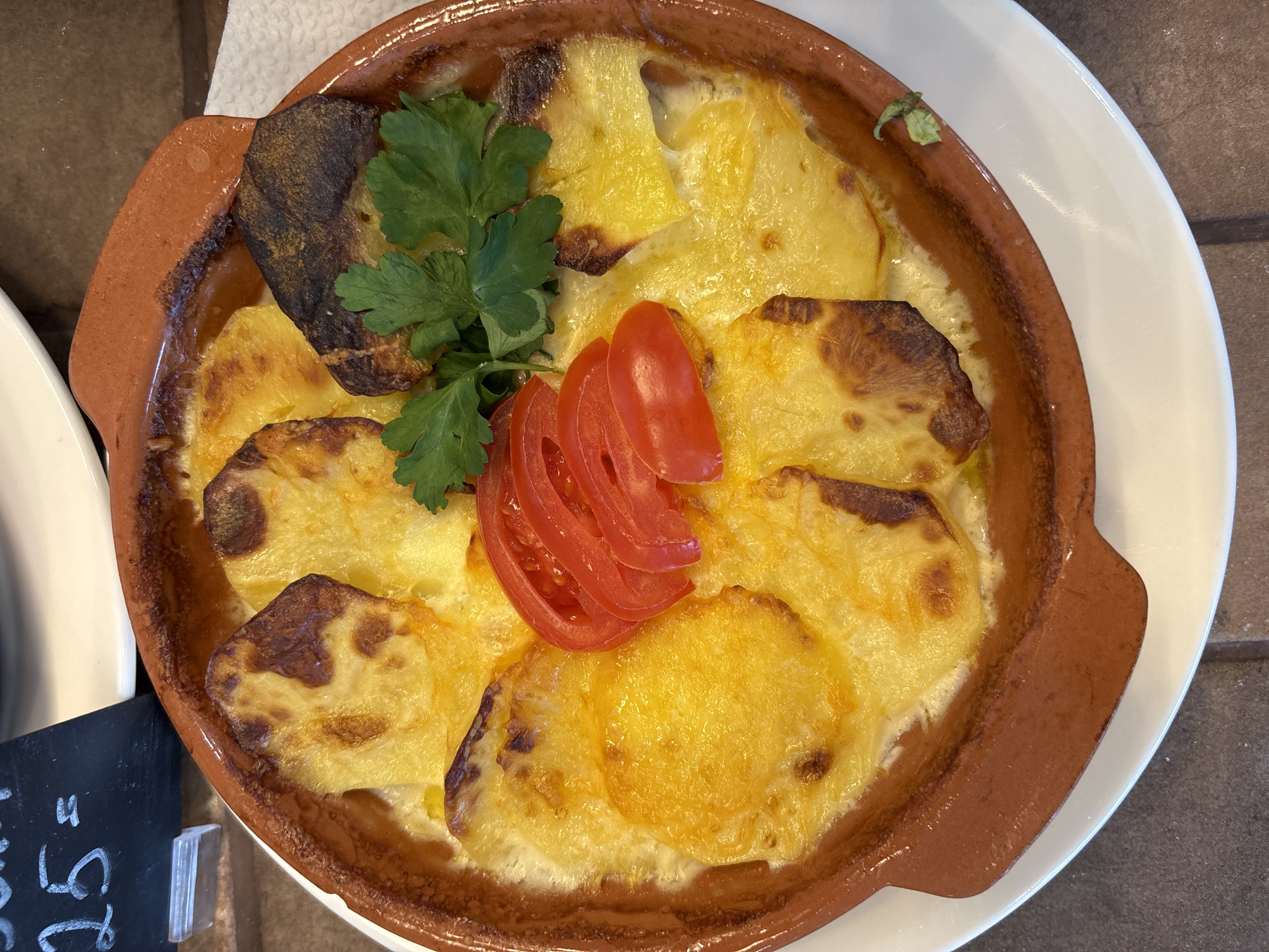 The image shows a close-up, overhead shot of a dish of baked potatoes au gratin served in a terracotta bowl. The potatoes are sliced and browned on top, with visible cheese sauce bubbling around them. The dish is garnished with fresh parsley and thinly sliced tomato arranged in the center. The terracotta bowl sits on a white plate, which is placed on a dark brown surface, possibly a wooden table. To the lower left of the bowl, a small black sign displays the price "25", likely referring to the cost of the dish. The setting appears to be in a restaurant setting, although there's no visible information to confirm the city. Therefore, it is not possible to confirm the location in Izhevsk, Russia with the information available in this image.
