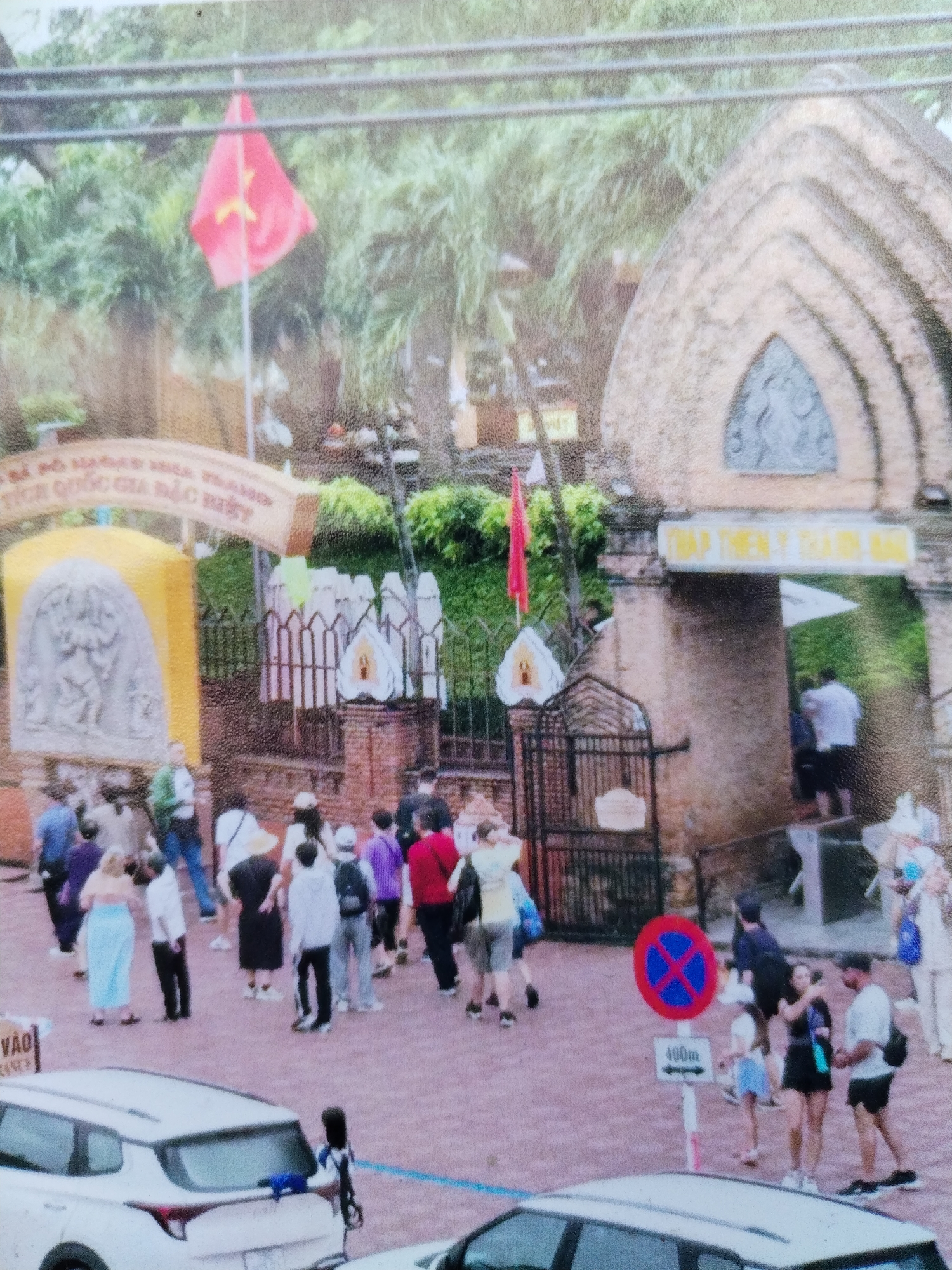 The image depicts a crowded entrance area, likely a tourist attraction, located in Bac Nha Trang Ward, Vietnam. A group of people is milling around, some entering through a gated archway, others standing on the paved plaza in front. At least two cars are parked near the entrance.
The entrance is adorned with brick structures and decorative elements, including a yellow sign with carvings. Two flags are visible: one, a Vietnamese flag on the left, and another near the right archway. A "no parking" sign is visible near the right side of the plaza. The environment is lush with greenery in the background.
