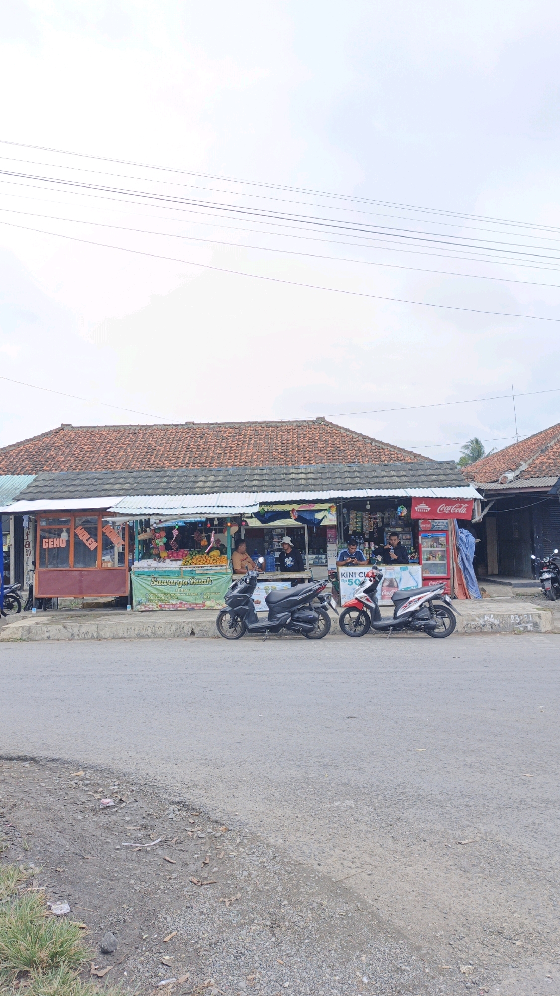 This is a full-shot, eye-level, outdoor photograph taken in Desa Karangbenda, Indonesia. The image features a roadside scene with a small commercial building or stalls situated along a paved road. The building has a tiled, orange-brown roof supported by a white metal structure. In front of the building, there are several stalls selling goods, including what appears to be fruit, displayed in baskets and on tables. A red Coca-Cola refrigerator is visible on the right side.
Three motorcycles are parked in front of the stalls. Two are in the foreground, a black scooter on the left and a red and white scooter on the right. A third motorcycle, partially visible, is parked further down the road on the right.
Several individuals are present in the scene. Two men are sitting at a table in front of one of the stalls, seemingly interacting or taking a break. Another man is standing behind a counter, possibly attending to customers. On the far right, a person is standing near the Coca-Cola refrigerator, and another person is partially visible exiting a stall on the left.
The environment is somewhat rustic, with the paved road showing some wear and tear. The ground beside the road is a mixture of dirt, gravel, and scattered leaves. Overhead, power lines stretch across the frame. The sky is overcast with some hints of blue. The overall impression is a typical street-side scene in a rural or semi-urban Indonesian setting.