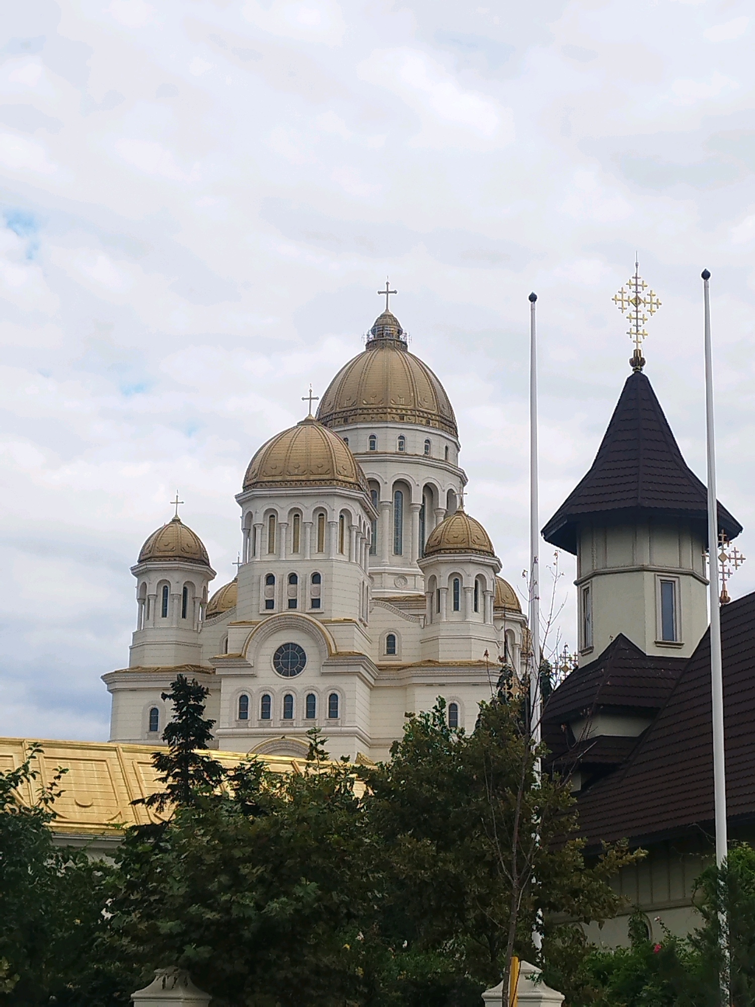 The image shows a large, white Orthodox cathedral with golden domes in Bucharest, Romania. The cathedral is a prominent landmark and features multiple domes of varying sizes, topped with crosses. In the foreground, there is greenery, including trees, and a glimpse of another building with a dark, sloping roof. Two tall, thin poles with small flag-like tops stand in front of the cathedral, and another decorative golden cross is visible atop the smaller building. The sky is overcast with clouds. The cathedral is the "Catedrala Mântuirii Neamului Românesc" or the National Salvation Cathedral.