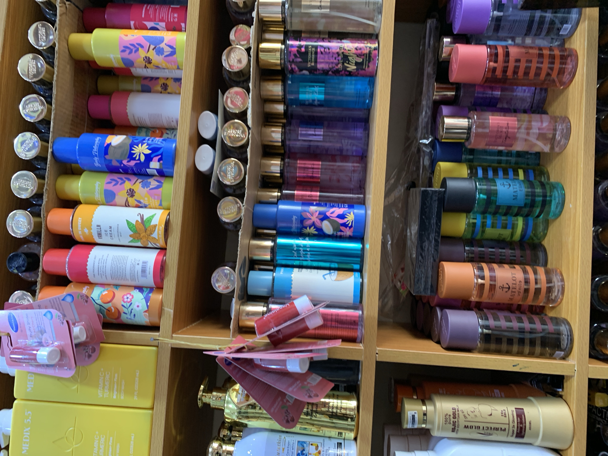 The image shows a shelf stocked with various personal care and beauty products, likely in a retail setting. The shelves are made of light brown wood. The products include body washes, lotions, lip balms, and fragrance sprays in different colored bottles and packaging. Brands such as "Medix 5.5", "Perfect Glow", "Philosophy", and "Vaseline" are visible. The bottles are stacked horizontally, with labels facing forward. Some products appear to be individually packaged, while others are grouped together. There is no visible indication of the location being Jos, Nigeria.