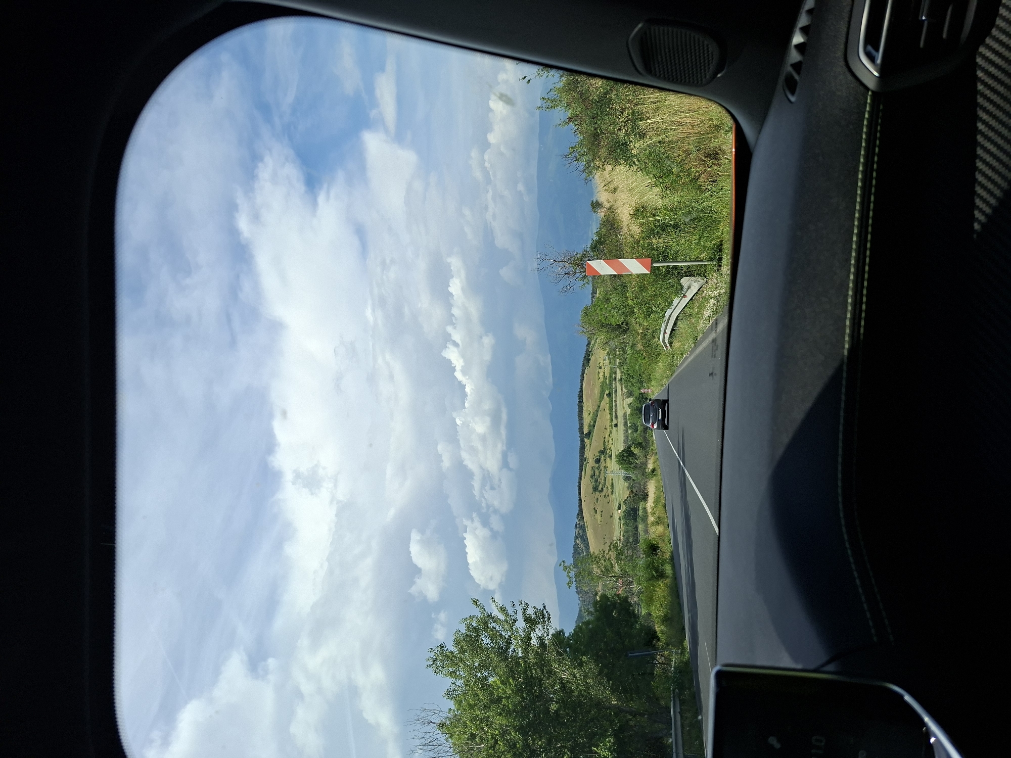 The image captures a panoramic view from inside a vehicle, looking out through the front windshield onto a scenic landscape in Mihiltsi, Bulgaria. The road, a two-lane asphalt path with a solid white dividing line, curves gently ahead. In the distance, another black car, possibly a 4x4 vehicle (faintly visible "4X4" on its rear), is driving in the same direction.
The setting is characterized by rolling green hills and scattered trees, suggesting a rural or mountainous region. A vast body of water, likely a lake, stretches across the mid-ground, nestled between the hills and extending towards the horizon. The sky above is bright blue with scattered white cumulus clouds.
On the right side of the road, a distinctive red and white striped warning sign, possibly indicating a sharp curve or hazard, stands prominently. A metal guardrail runs along the roadside. The interior of the car frames the view, with the dashboard, a speaker grille on the left, an air vent, and possibly carbon fiber trim on the right, all in dark tones. Green stitching can be seen on a dark upholstered surface on the right, adding a subtle detail to the vehicle's interior. A faint reflection of a digital display, showing "1.0", is visible on the bottom right of the image, possibly from the car's console.
The scene depicts a daytime journey under clear, sunny weather, highlighting the natural beauty of the Bulgarian landscape.