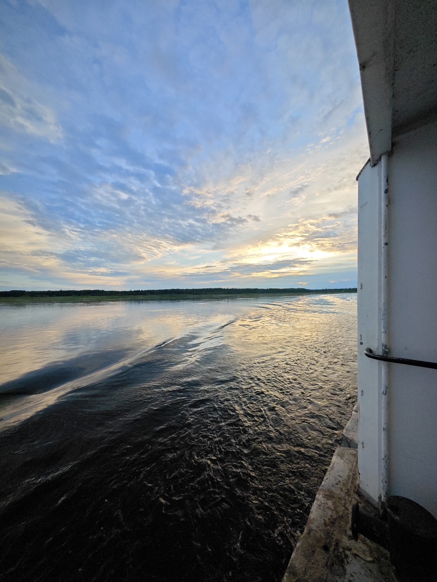 The image depicts a serene river scene at sunset, viewed from the side of a vessel. The sky is adorned with wispy clouds, catching the golden hues of the setting sun, with patches of pale blue still visible. The water, reflecting the sky's colors, appears dark and rippled, with a prominent wake trailing from the viewer's perspective. A distant, treed shoreline forms a dark horizontal line across the horizon. On the right side of the frame, a white, weathered wall of the vessel is visible, with a dark railing attached. The concrete edge of the vessel's deck is also present in the foreground. The scene evokes a sense of peaceful travel and the beauty of nature at dusk. The time of day is clearly sunset. The weather appears calm and pleasant. The location is implied to be a river in Nikulino, Russia, based on the context provided. There is no visible text in the image.