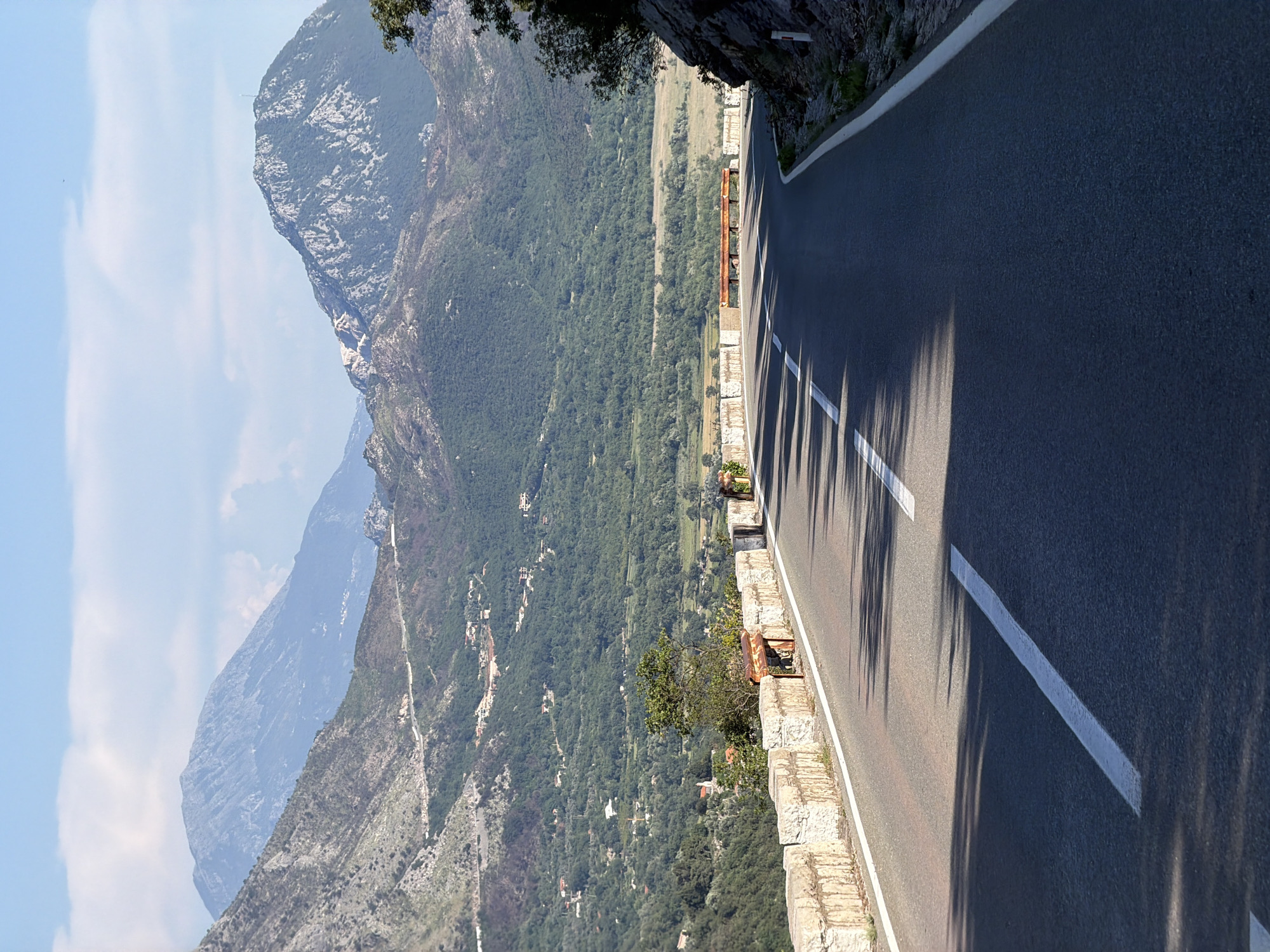 The image depicts a mountain road near Androvići, Montenegro. The road is narrow, with a concrete barrier on one side protecting a steep drop into a forested valley. A mountain range is visible in the distance, partially obscured by haze. The road surface is asphalt with white line markings. Tree shadows are cast onto the road.