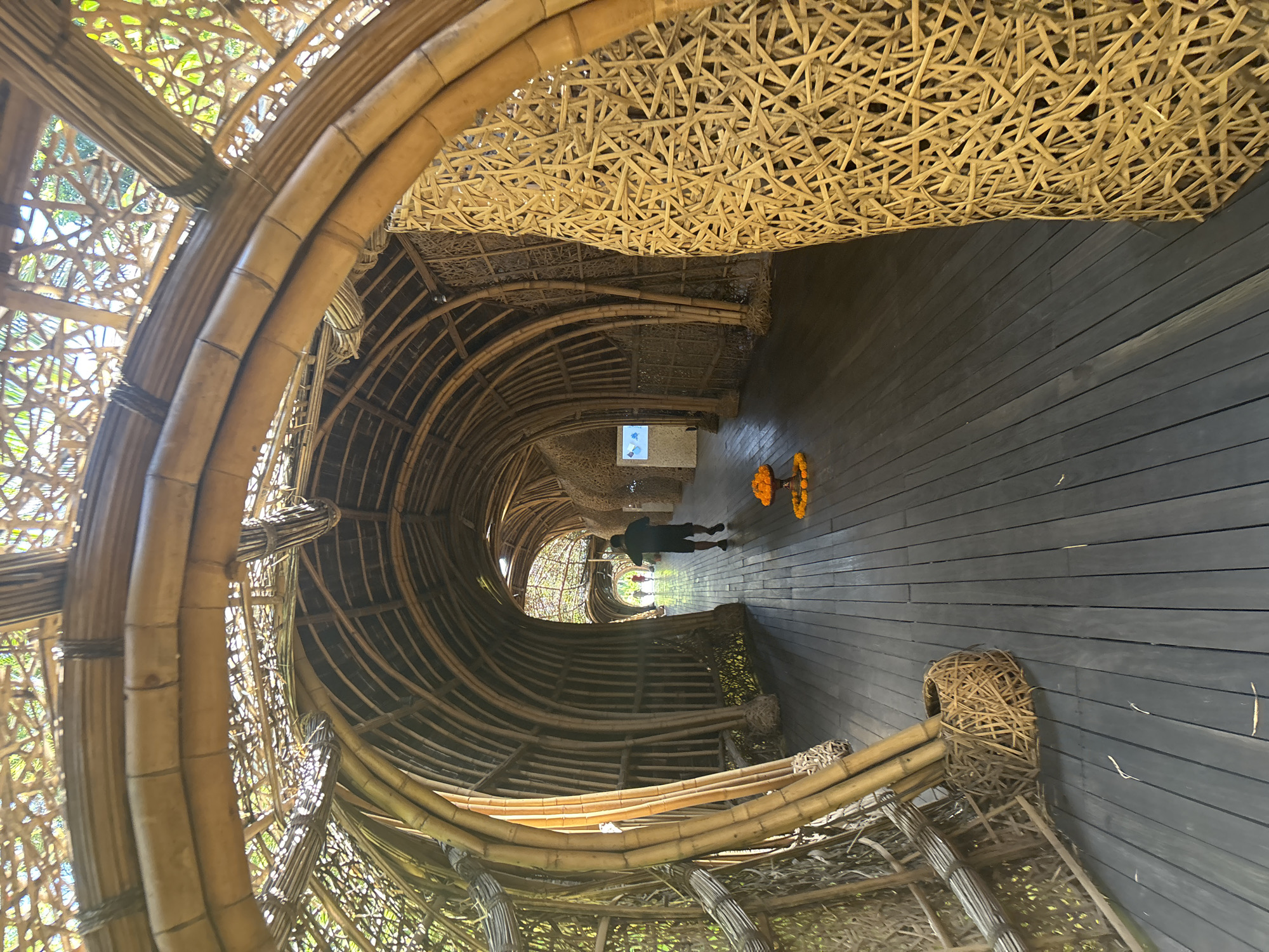 This is a wide-angle, eye-level, outdoor shot of a bamboo structure in North Kuta, Indonesia. The structure forms a long, curved tunnel with a wooden floor. The tunnel's walls and ceiling are constructed from numerous bamboo poles of varying thicknesses, woven together in an intricate lattice pattern. Light filters through the bamboo, creating a dappled effect on the floor.
In the foreground, to the left, is a basket-like structure made of woven bamboo, holding several upright bamboo poles. Further down the tunnel, a person is walking away from the camera, wearing a dark t-shirt and shorts. In front of this person, on the wooden floor, is a small, circular arrangement of orange flowers.
The tunnel recedes into the distance, with more of the bamboo lattice structure visible. On the left wall, a small digital display screen is mounted. The overall impression is one of organic architecture and a natural, tranquil environment.