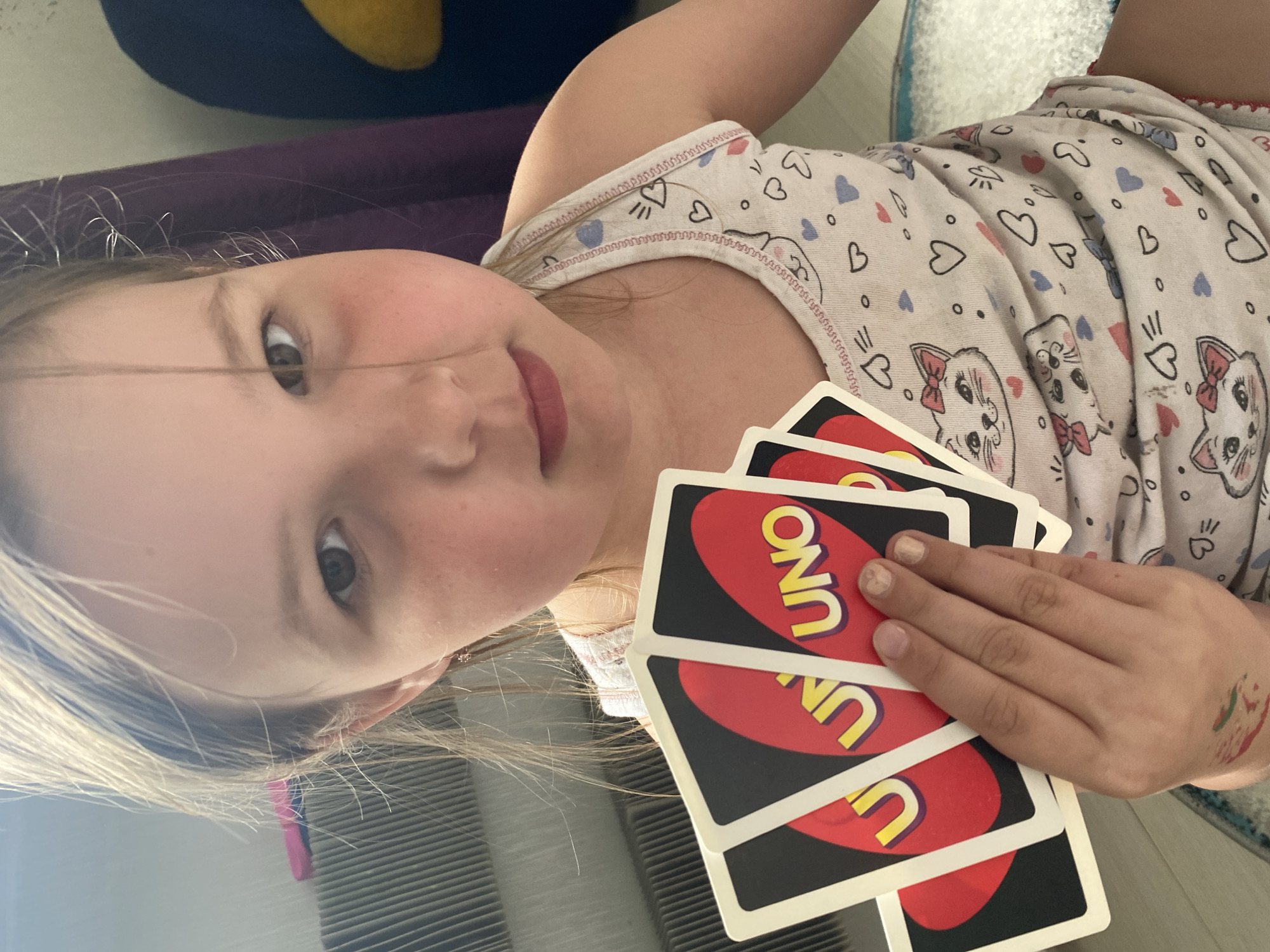 This image depicts a young Caucasian female holding a hand of UNO cards in an indoor setting, identified as the city of Chaikovsky, Russia. The girl, appearing to be a child or pre-teen, has light brown hair, fair skin with noticeable flush on her cheeks, and brown eyes directly engaging the viewer. Her lips are closed in a neutral expression.
She is dressed in a light-colored tank top adorned with a repeating pattern of cartoon cat faces, red and blue hearts, and small dots, featuring red stitching along the trim. Her left hand, positioned centrally in the frame, holds a fan of at least five UNO cards, predominantly red with yellow "UNO" branding. Her fingernails show signs of chipped light-colored polish. A small, multi-colored temporary tattoo or mark, consisting of red, green, and blue streaks, is present on her left forearm. A small, simple gold-colored earring is visible in her right ear.
The background on the left reveals a light-toned, possibly ribbed or slatted surface, suggestive of furniture or a radiator cover, with a partial pink object visible behind it. To the right, dark purple fabric, potentially a curtain or blanket, is visible, partially obscuring a dark blue object with a yellow curve. A light-colored, textured rug or carpet is partially visible in the lower foreground. The overall scene is casually lit, consistent with an interior residential space.