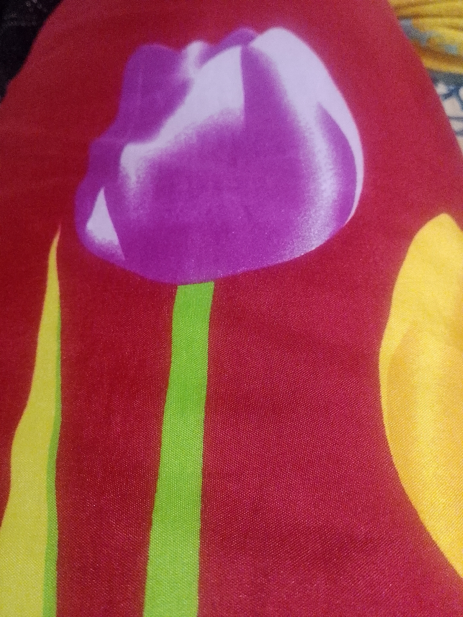 The image shows a close-up of a patterned fabric. The background is a deep red color. A large, stylized tulip design is prominent. The tulip's flower is primarily light purple with some white shading. The stem is green. A portion of a yellow shape, potentially another petal or leaf, is visible on the right side of the frame. The fabric's texture is visible, with a woven pattern discernible. There is no indication of the city of Lhokseumawe, Indonesia, or people or interactions within the frame.