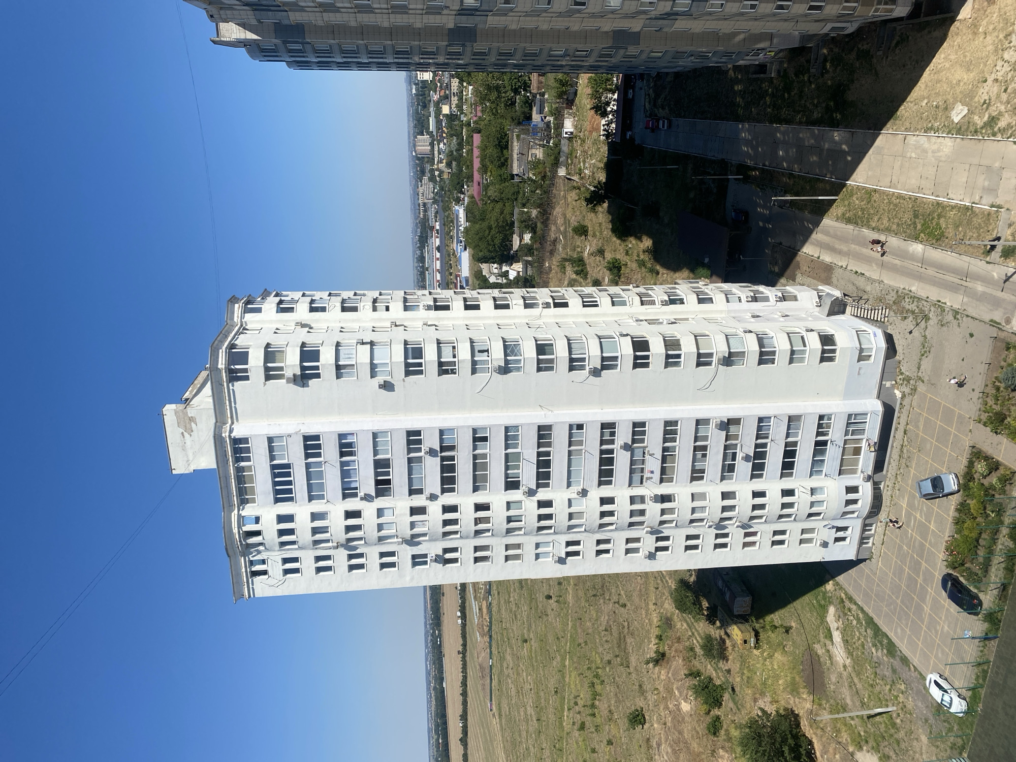The image depicts a view from a high vantage point overlooking an urban area on a clear, sunny day. In the foreground, a tall, white apartment building dominates the scene, characterized by its numerous windows and multiple stories. To the right, another apartment building, this one grey with a patterned facade, is partially visible. Between the two buildings, a paved road or walkway stretches into the distance, casting long shadows from the buildings. Several cars are parked along the side of the road and in a courtyard area in front of the white building, where a few individuals can be seen walking. Beyond the buildings, a flat, grassy field with some scattered vegetation extends towards a more developed urban landscape with smaller buildings, trees, and more distant structures. The sky is a deep, cloudless blue. The scene appears to be in the city of Chornomorsk, Ukraine, given the typical architecture and landscape for the region.