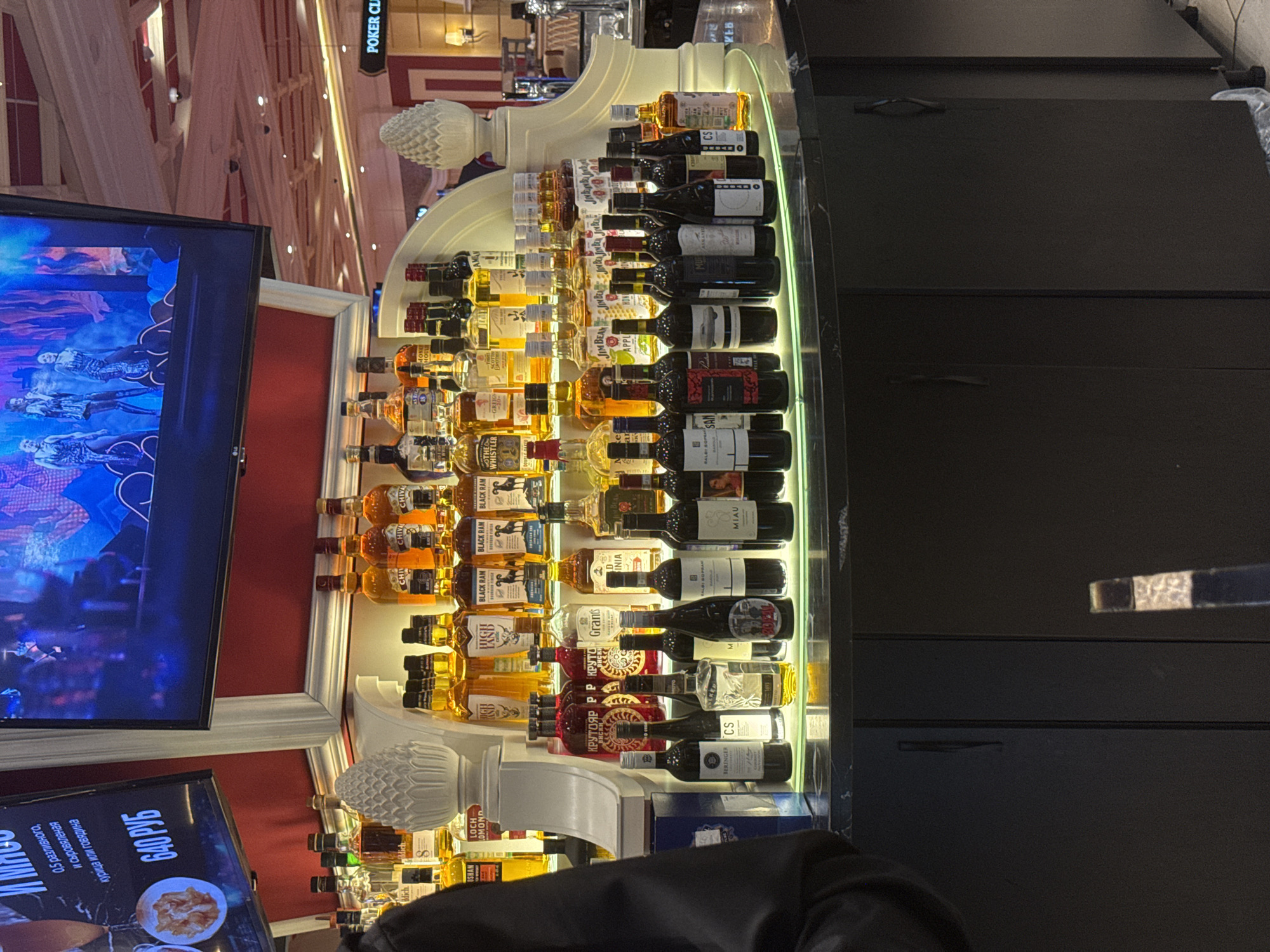 The image captures a bar setting, likely in Shkotovo, Russia, as indicated by the Russian text on a nearby screen displaying "640 РУБ", which translates to "640 Rubles", next to food images. The bar features a lavish display of various liquor and wine bottles arranged on illuminated shelves behind the counter. Brands like Chivas, Black Ram, The Whistler, and Jim Beam are visible, along with Russian labels such as “КРУТОЯР” (Krutoyar). A large TV screen is mounted nearby, showing a vivid, theatrical performance, potentially related to a show called "WEB". The bar is partially obscured by a dark-clad person, presumably a patron, and the countertop, made of what appears to be a sleek black material. The background shows ornate decorative elements and a sign saying "POKER CLUB".