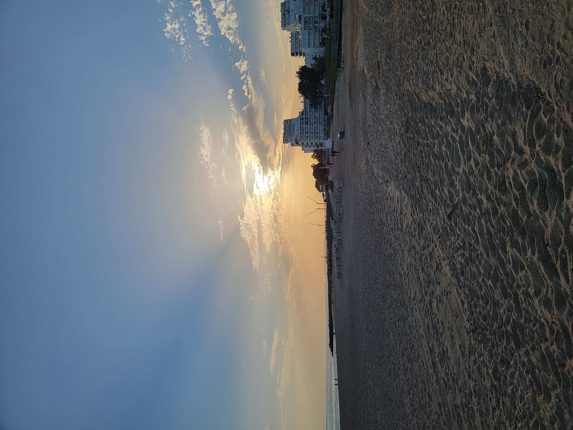 The image depicts a sandy beach in Quarteira, Portugal, during a sunset. The sun is low in the sky, casting a warm glow and creating visible light rays through the scattered clouds. The foreground is dominated by the textured sand of the beach, with subtle patterns from wind or previous foot traffic.
In the middle ground, there are rows of empty lounge chairs and umbrellas set up, indicating a tourist area. Beyond the chairs, a walkway and some vegetation can be seen. Several buildings, appearing to be hotels or apartment complexes, are visible in the background, characterized by their white facades and multi-story architecture. There are a few people walking along the beach and near the buildings. The ocean stretches to the left of the frame, with a breakwater or pier extending into the water. The overall scene conveys a sense of tranquility as the day ends.